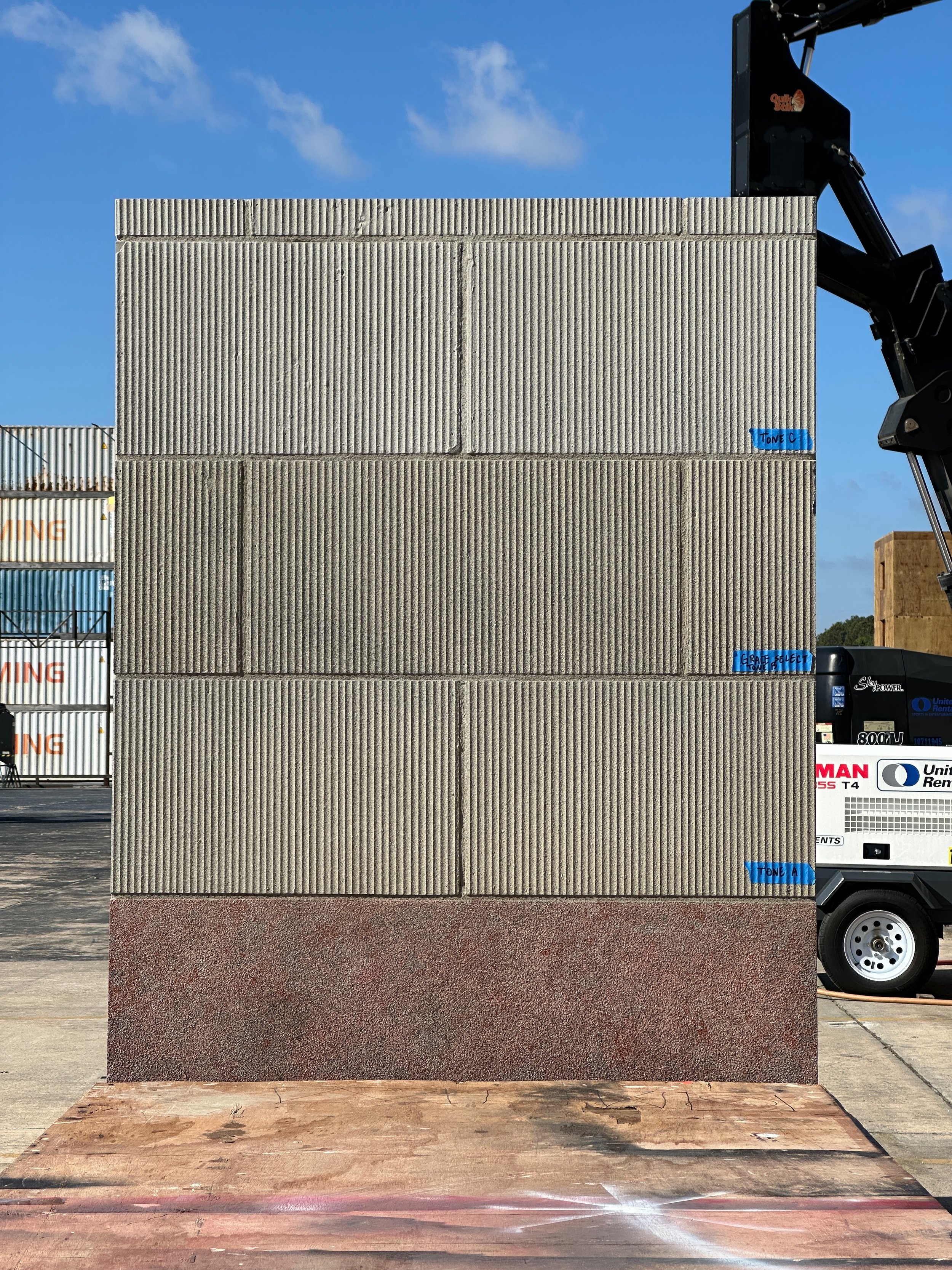 Stack of three different textured concrete blocks with blue tape labels, in an outdoor industrial area with a clear sky.