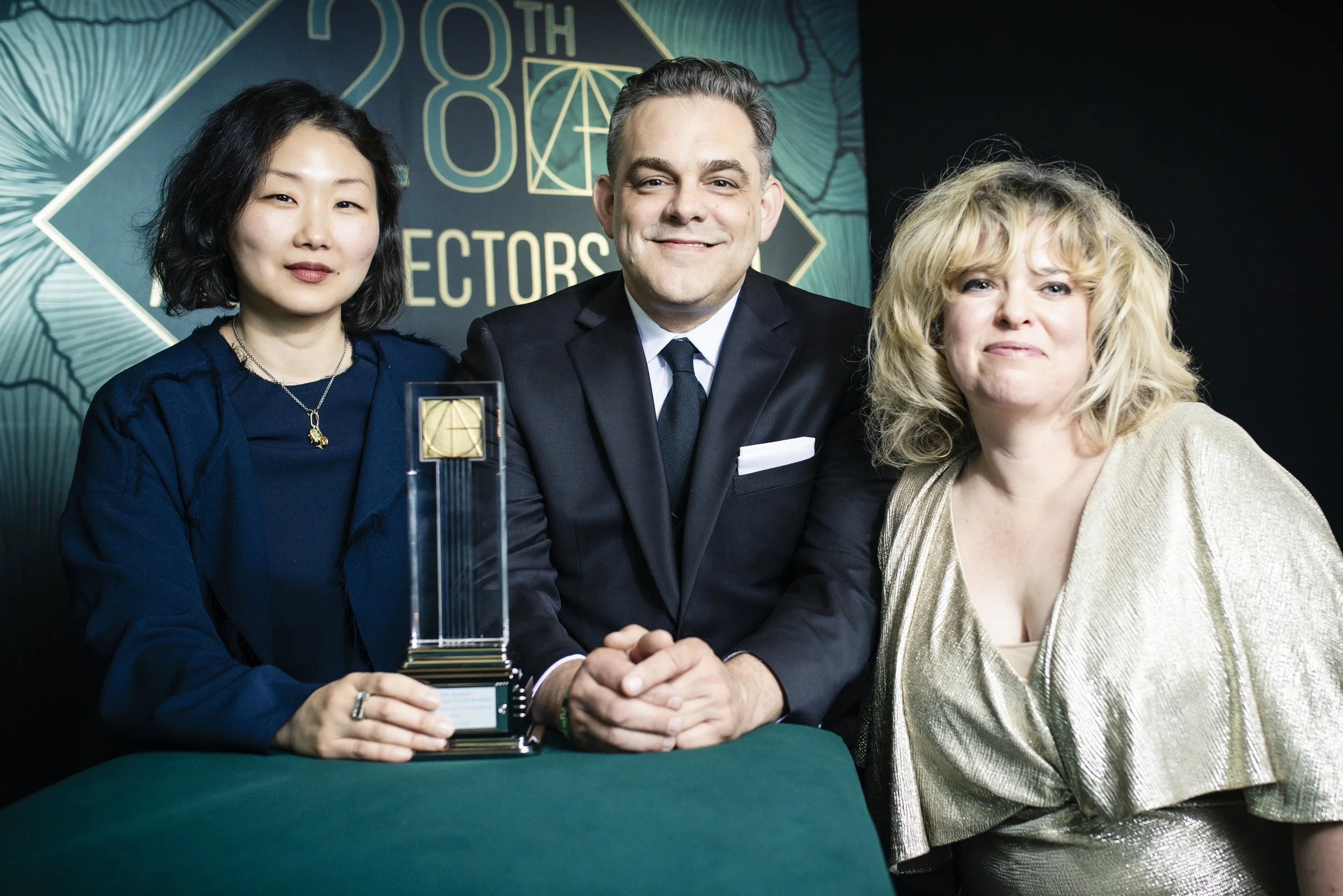 Three people sitting at a table during an awards ceremony, with a woman on the left holding a trophy, a man in the middle wearing a suit, and a woman on the right in a gold dress. A sign behind them reads '28th Editors'