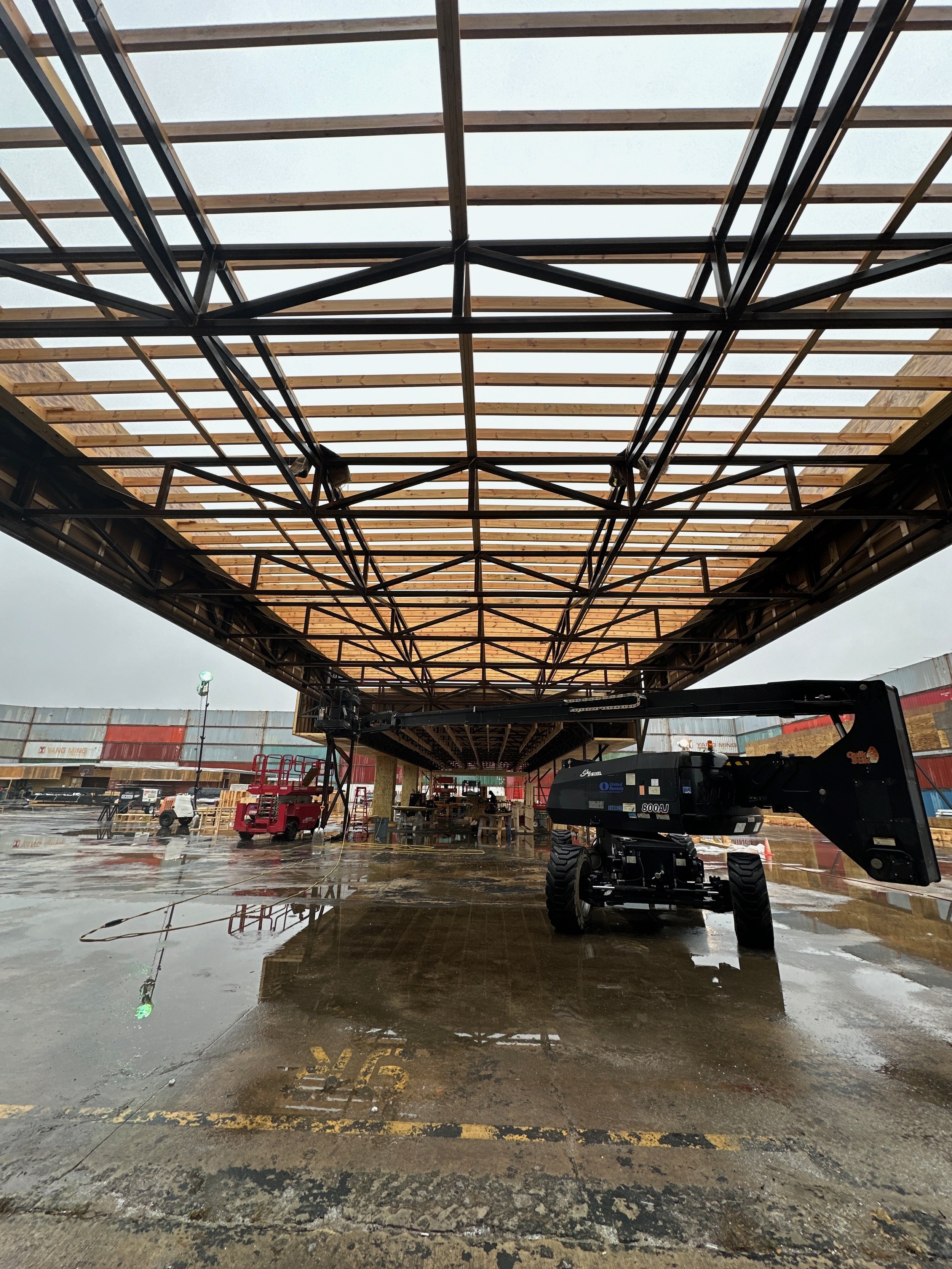 Construction site with a partially built roof supported by steel framework, construction equipment including a crane, and wet ground reflecting the structure, during overcast weather.