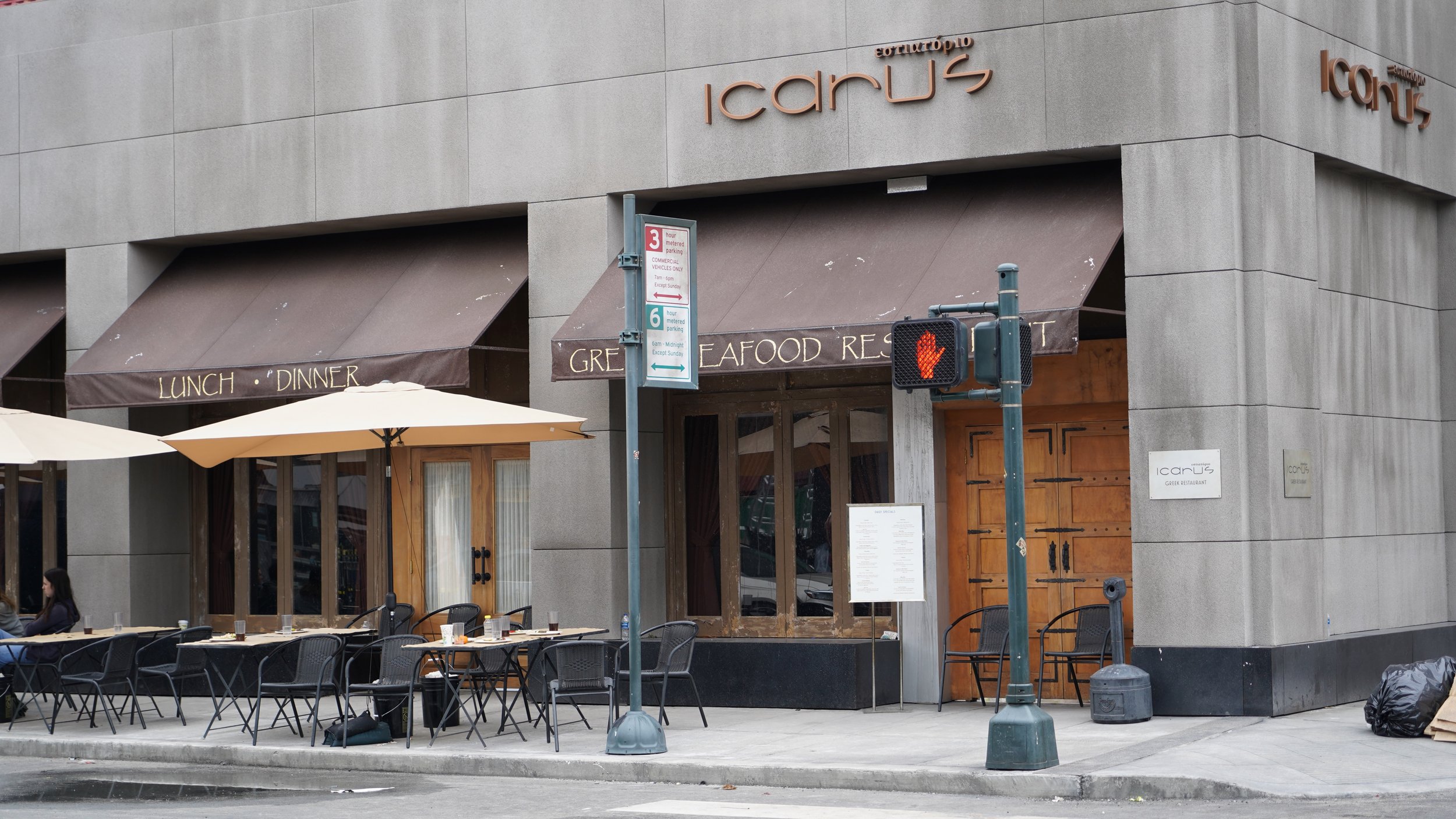 Exterior of a Greek restaurant named Icarus with outdoor dining area, tables, chairs, umbrellas, and a sidewalk with traffic signals.