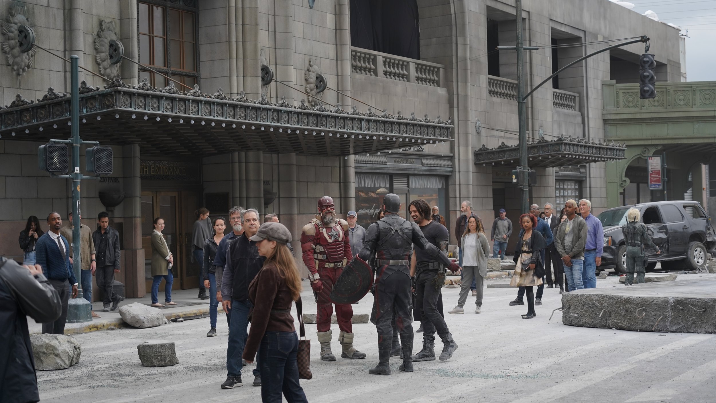 A film scene of a city street with many people, some dressed as superheroes, including one in red with a star emblem and another in black with a shield, with damaged cars, rocks, and debris on the ground.