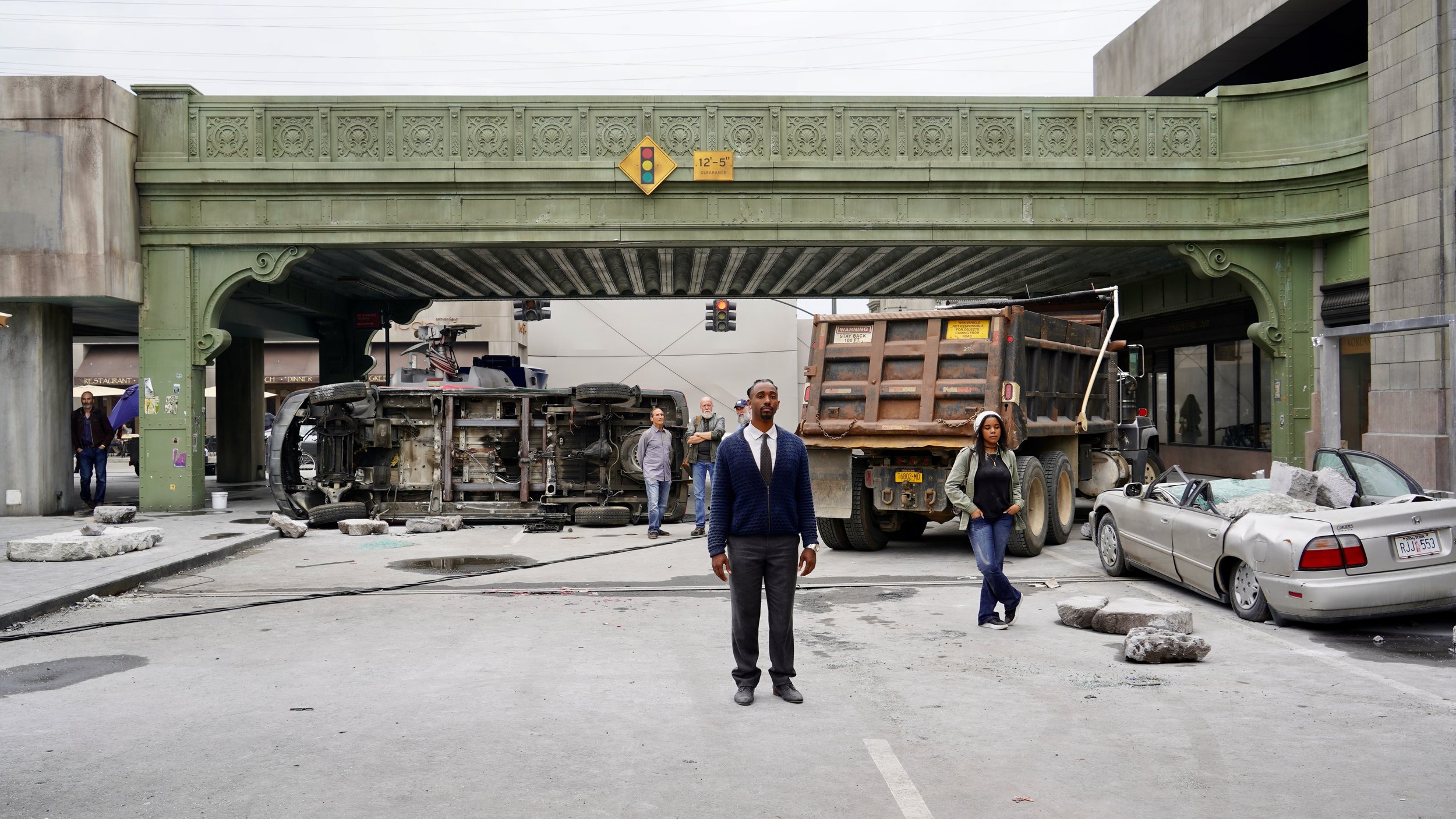 A man standing in front of a car accident scene under a bridge, with a flipped overturned vehicle, a large truck, and a damaged silver convertible car, while a woman and several people observe the scene.