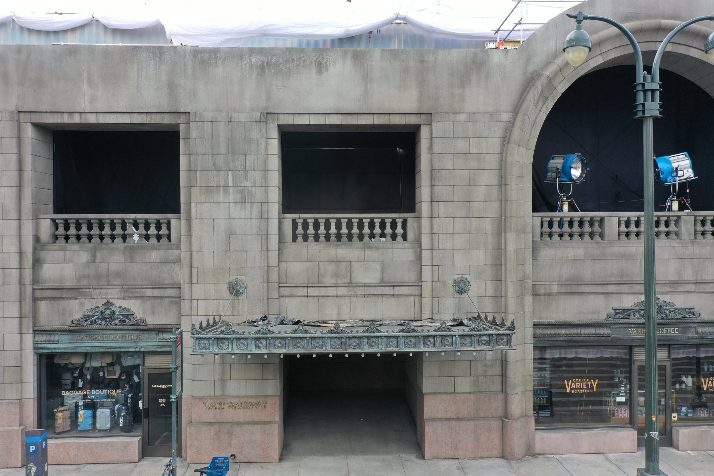 An unfinished building facade with large open sections and balconies, a decorative awning, and shop windows below. There are two large lights and a streetlamp in front.
