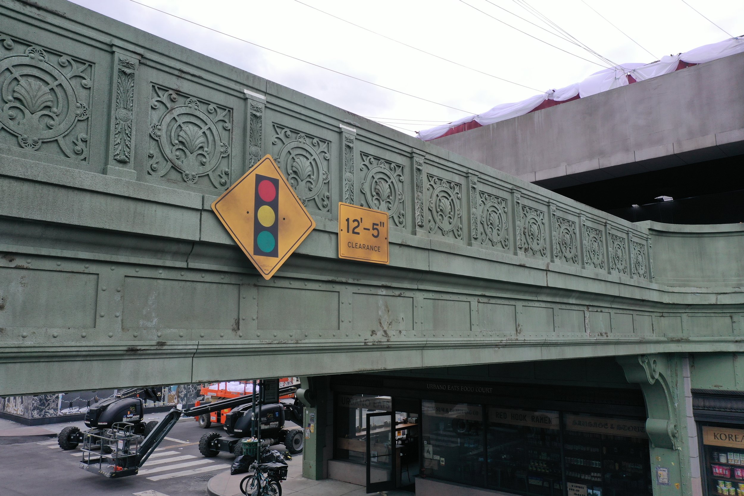 Close-up of a green overpass with decorative railings, traffic signals, and signs indicating a clearance of 12 feet 5 inches, underneath there is a street with parked vehicles, bicycles, and storefronts.