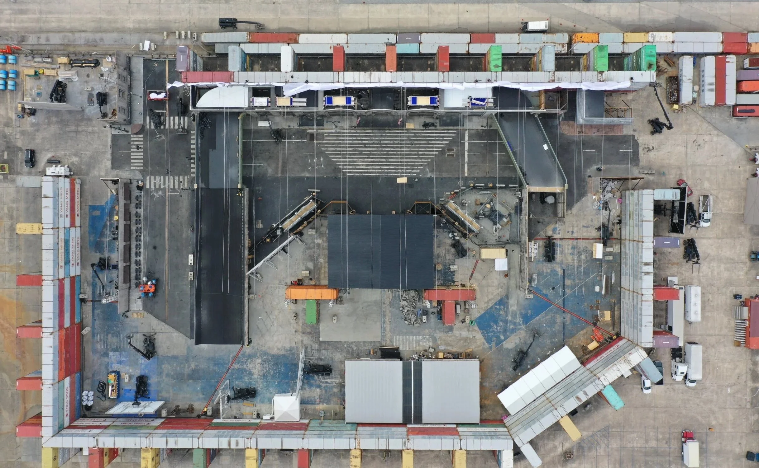An aerial view of an outdoor event stage setup in a large open area, surrounded by colorful barrier walls and various equipment, vehicles, and construction materials.