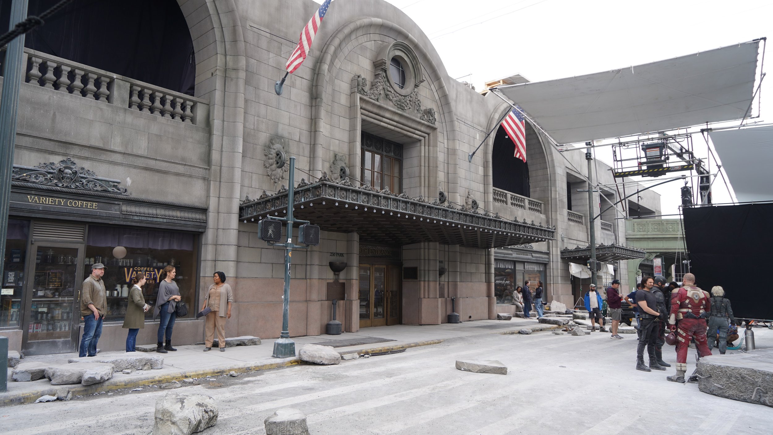 Behind the scenes on a film or TV set, showing a historic building facade with American flags, crew members, and equipment, including large black screens and scaffolding, with rocks scattered across the street.