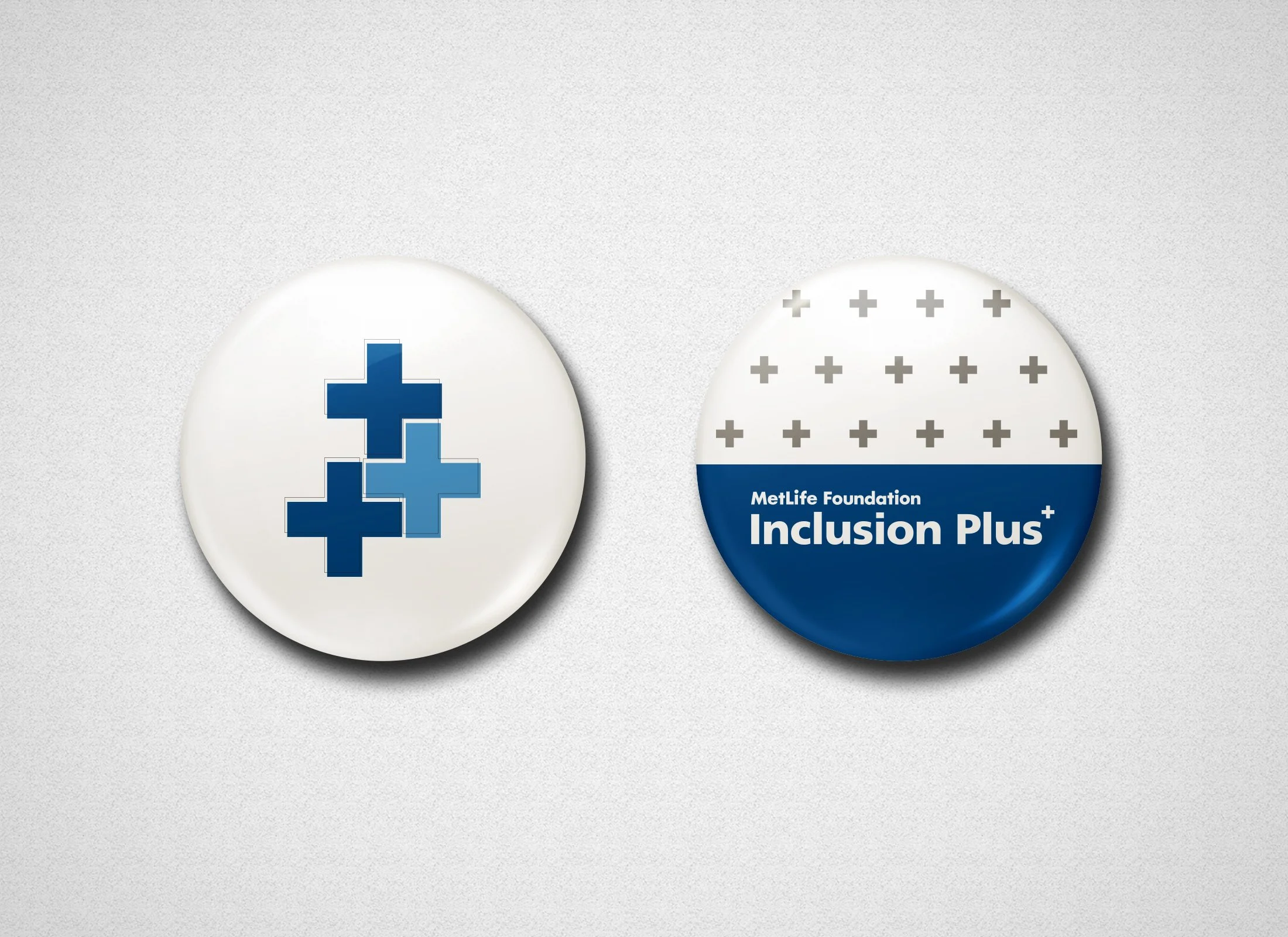 Two round badges, one with a blue cross composed of smaller blue puzzle pieces and the other with a pattern of gray plus signs and text that reads 'MetLife Foundation Inclusion Plus,' on a light gray textured background.