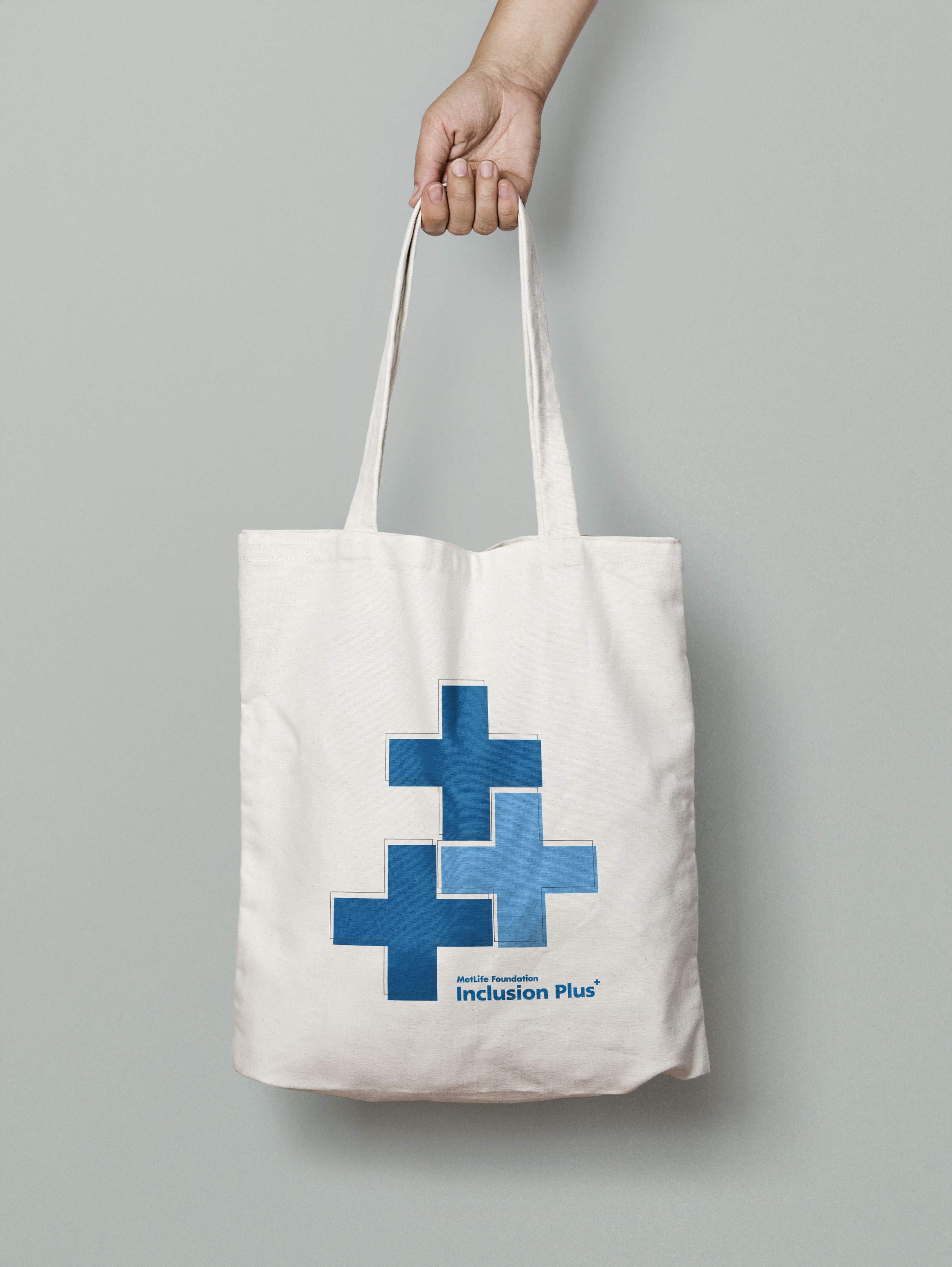 A hand holding a tote bag with a blue puzzle piece design and the text 'Inclusion Plus' from the MetaLife Foundation.