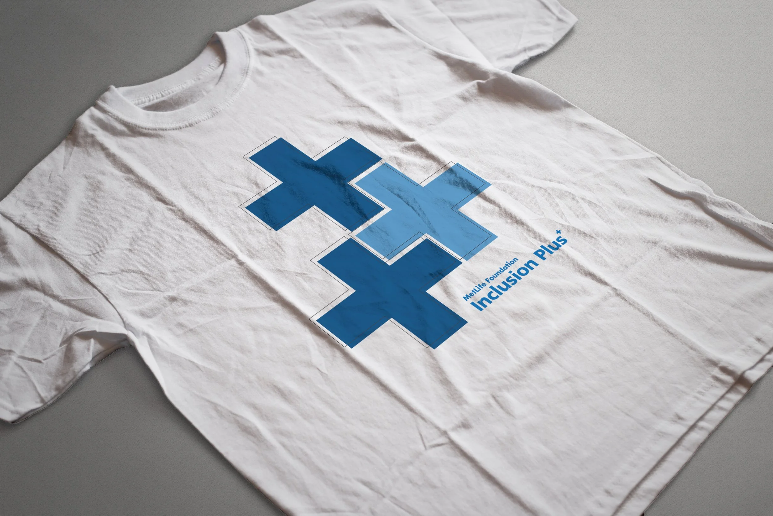 White T-shirt with blue logo and text for MetLife Foundation Inclusion Plus, wrinkled and laid flat on gray surface.