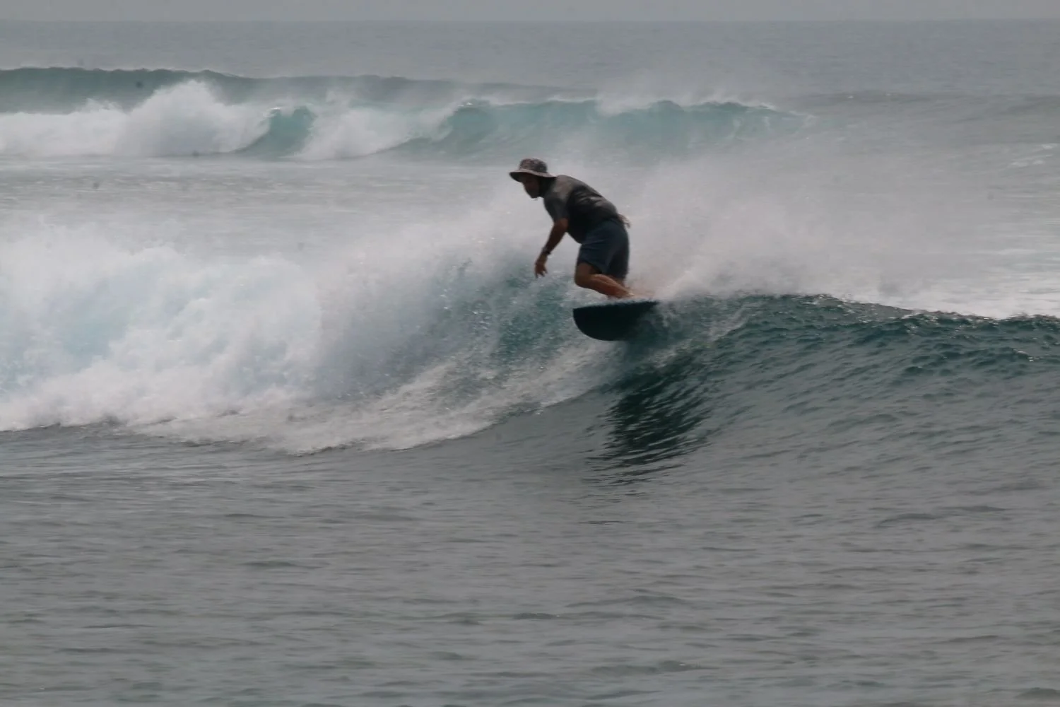 A person wearing a wide-brimmed hat and casual clothes surfing on a wave in the ocean.