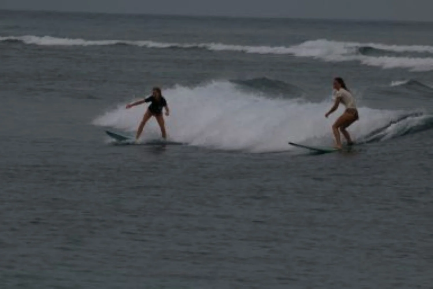 Two women surfing on the ocean with waves in the background.
