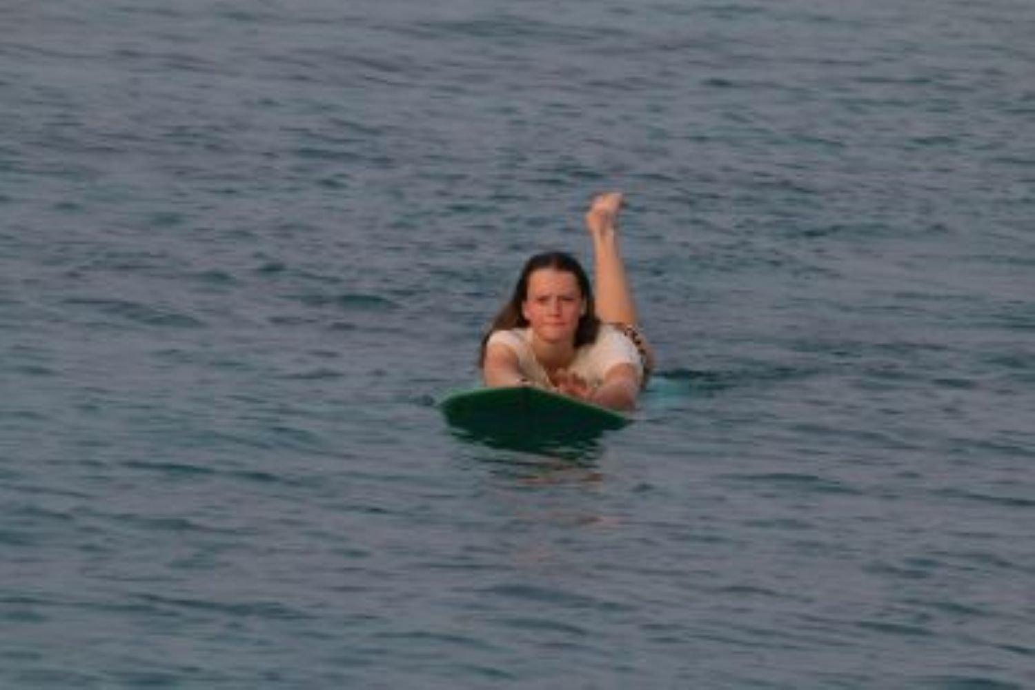 A woman lying on a surfboard in the water, reaching forward with her arms, with her head and one leg raised.