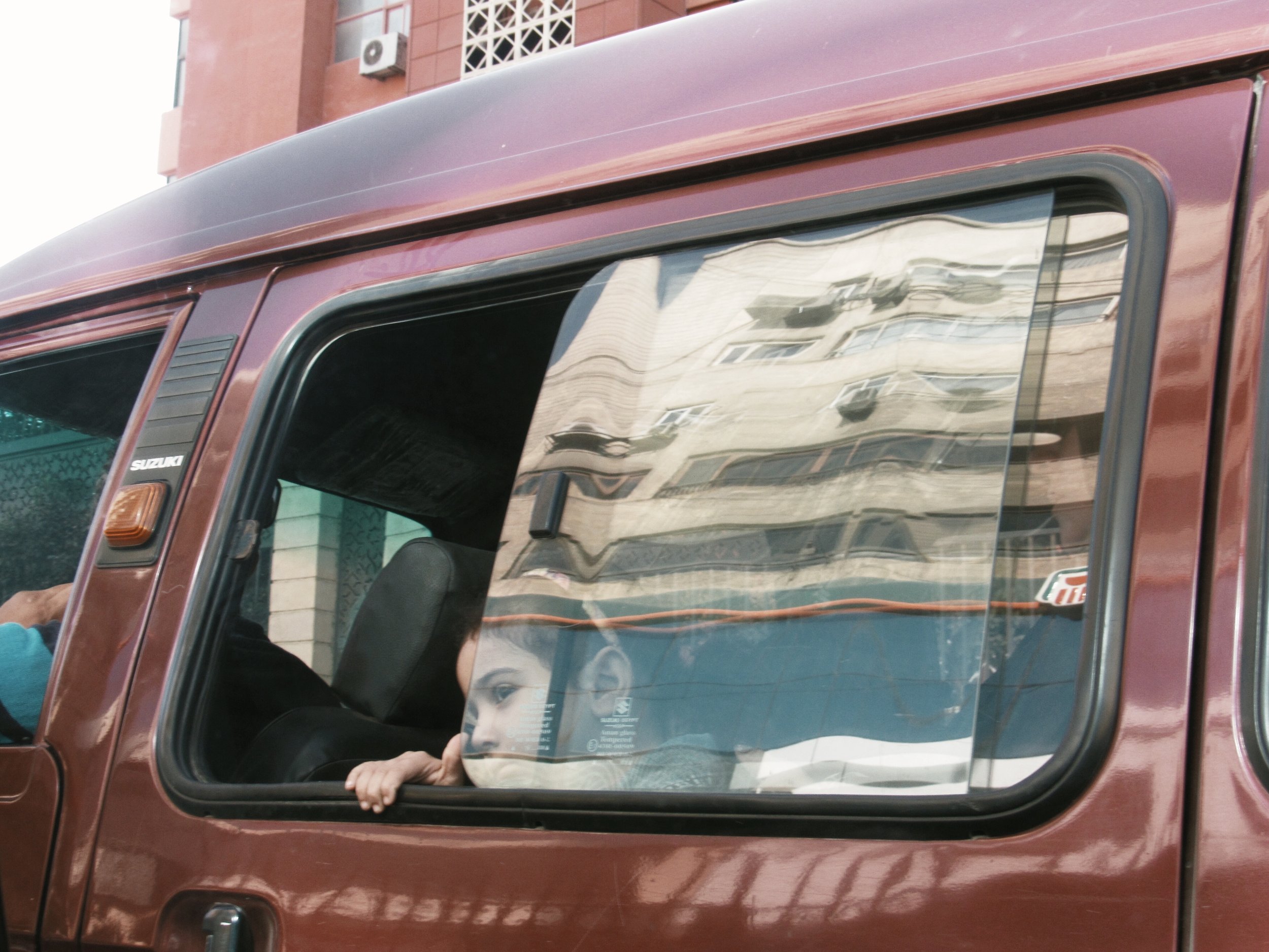 Girl in Traffic | December 17, 2018 Cairo, Egypt | A young girl with a wistful look upon her face peers her eyes out of a red micro-bus.