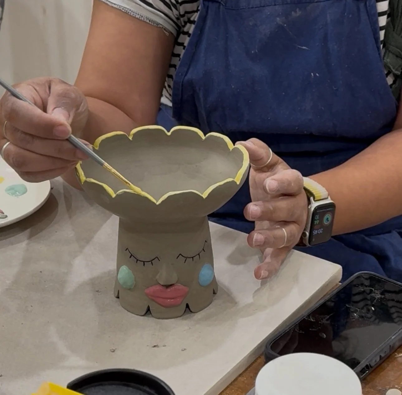 Person glazing a hand-built ceramic vase with face details during a pottery class