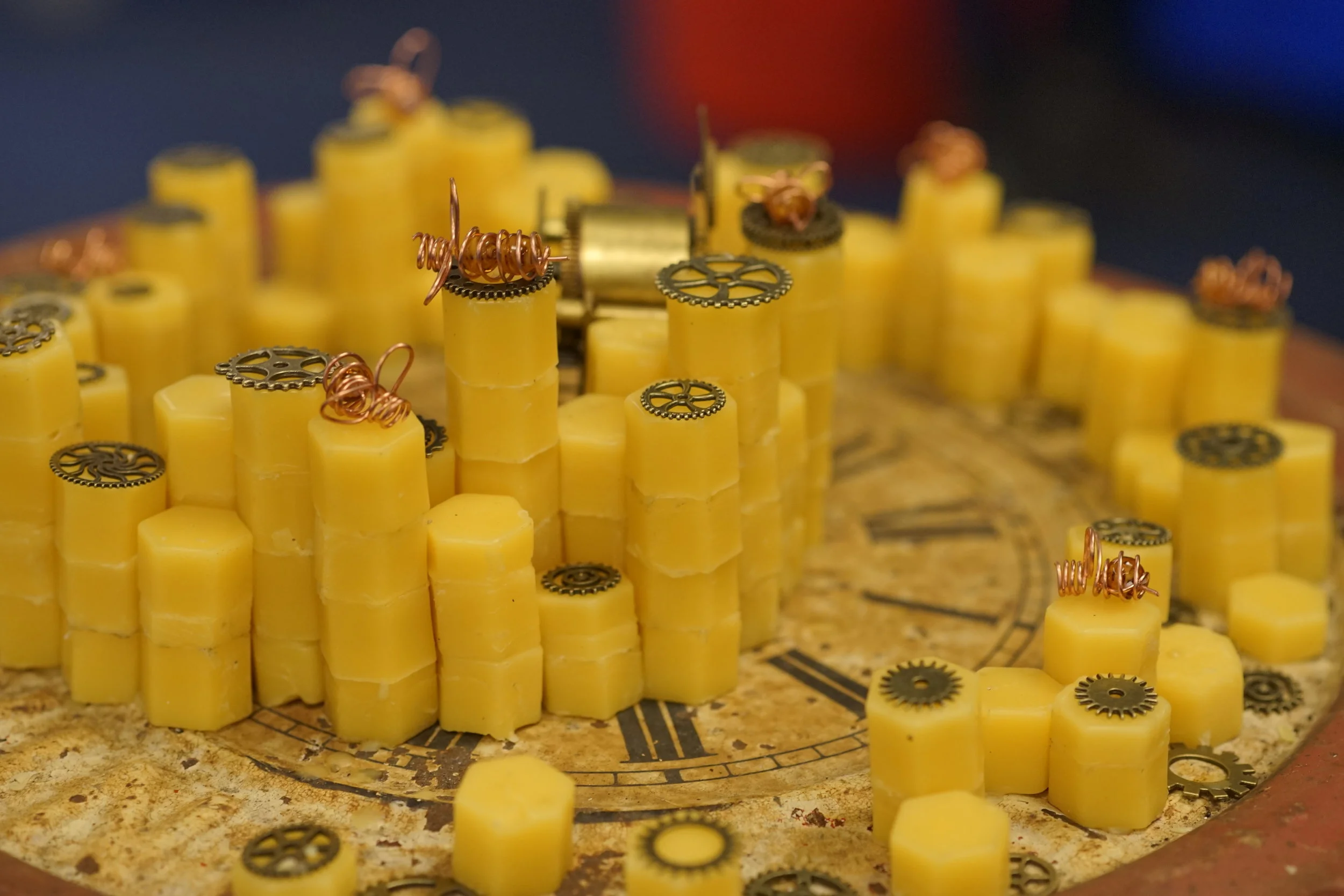 LAUREN NEVILLE - BEEHIVE COMPLEXITY THEORY Sculpture. Beehive Complexity Theory combines an antique clock and old parts with honeycomb shaped stacks of beeswax. The piece uses small copper wire renderings of bees to talk about the similarities between complexity theory in beehives and mechanics. 
