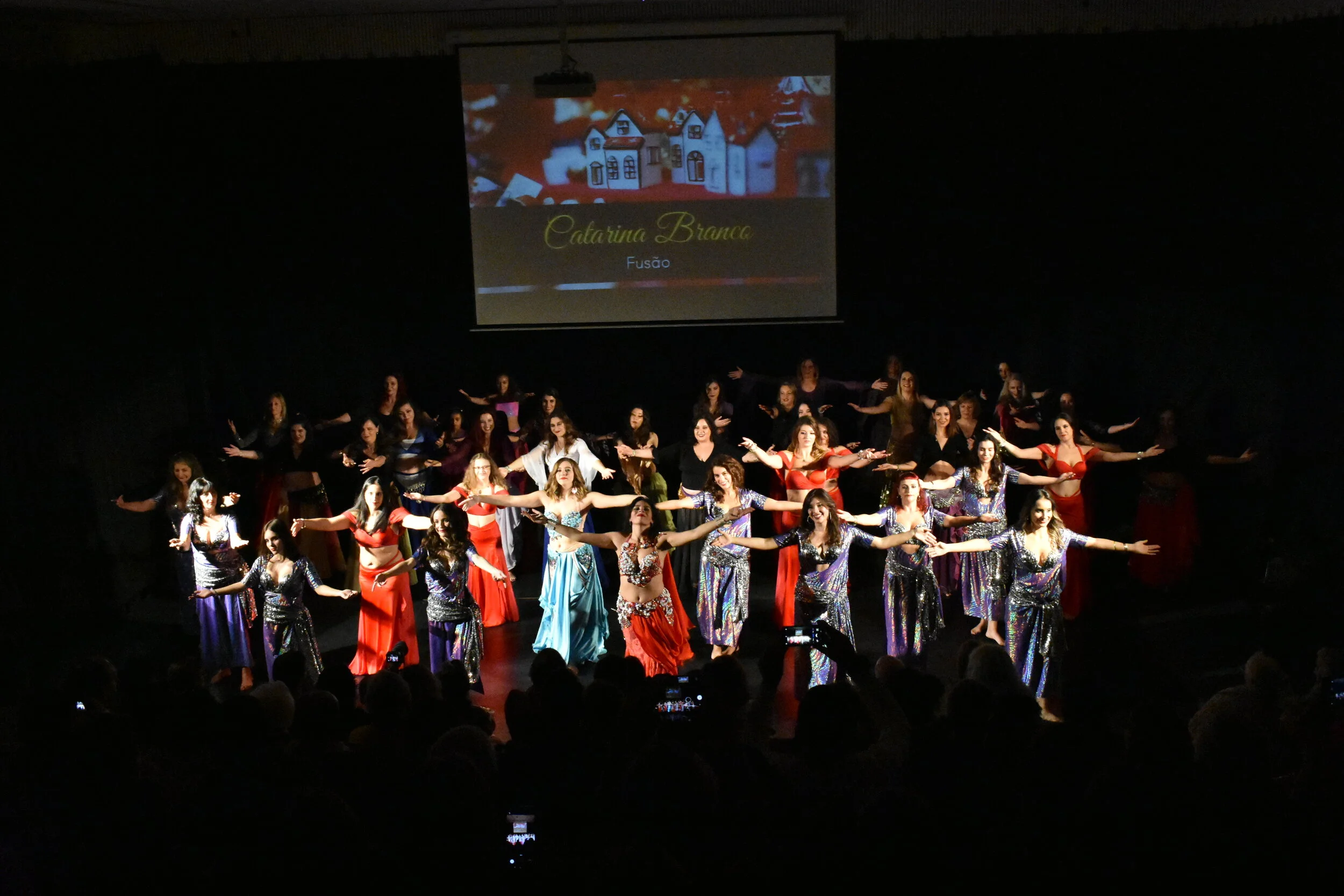 Grupo de pessoas em uma apresentação de dança no palco, usando roupas brilhantes, com um público assistindo ao fundo, e uma tela ao fundo exibindo nome "Catarina Branca" e a palavra "Fusão".