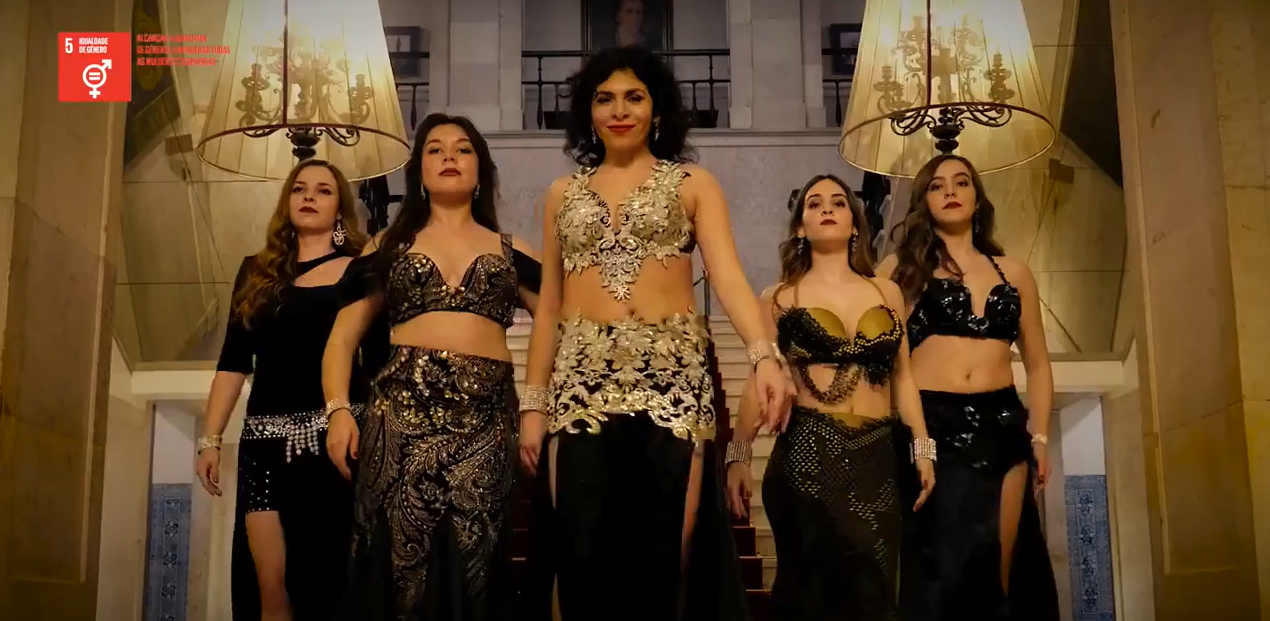 Mulheres vestidas com roupas sofisticadas descendo uma escada de borda dourada em um ambiente luxuoso