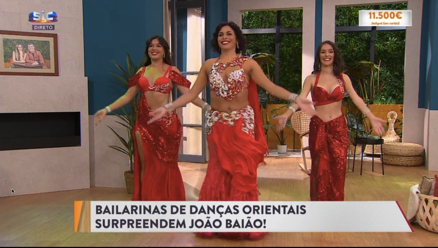 Três bailarinas de dança oriental vestidas com roupas vermelhas, realizando uma performance em um programa de televisão. Elas estão sorrindo e posando com os braços abertos. A cena é em um ambiente interno com decoração moderna e janelas de vidro com