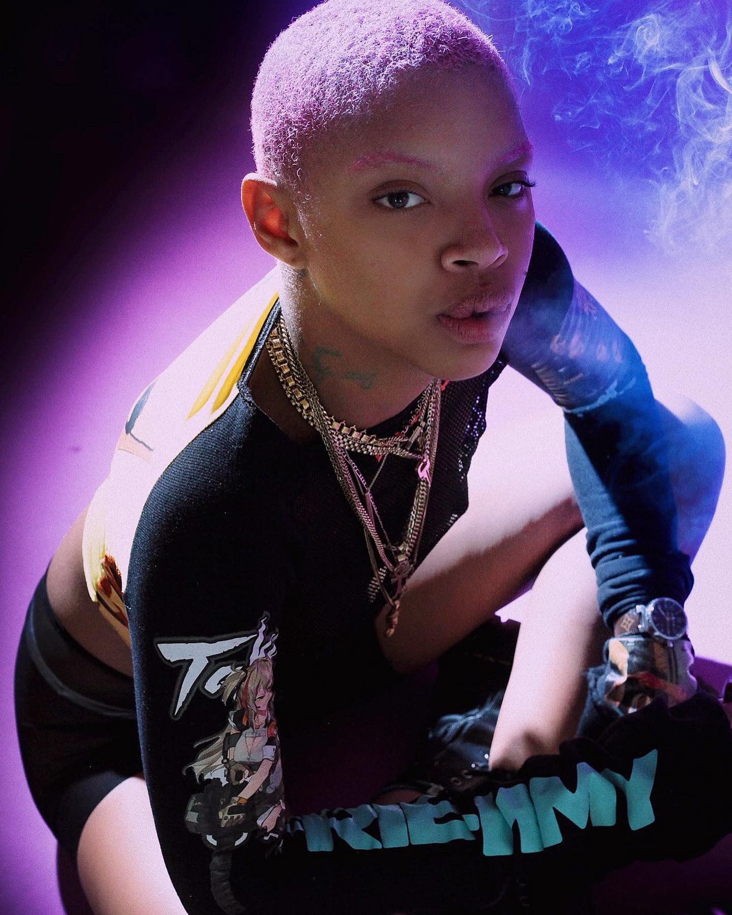 Young person with a short buzz cut hairstyle, wearing a black graphic jersey with colorful designs, layered necklaces, and a watch. They are leaning forward on a purple-lit floor with a smoky background.
