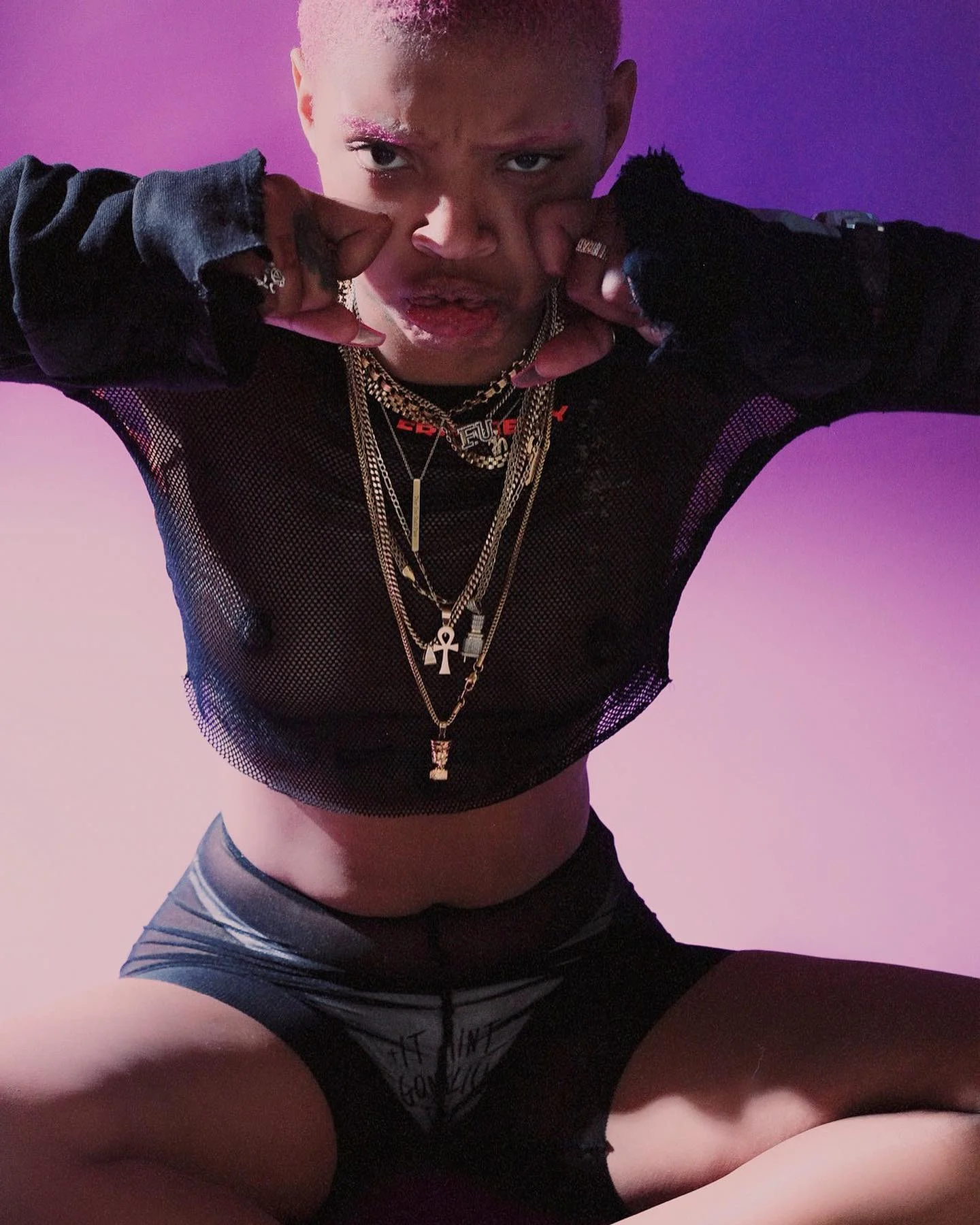 A person with short hair, wearing layered necklaces, a black mesh top, gloves, and shorts, making a fist gesture with a purple and pink gradient background.
