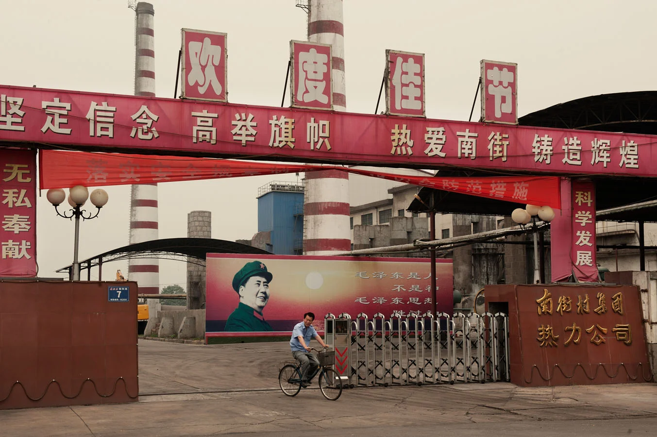 A worker leaving Nanjie heating factory on his bike. Above him a propaganda banner says: "Forge one's determination, hold the flag high, love nanjie and build a place of glory". Behind him, inside the factory a billboard featuring Mao with the writing"Mao Zedong is a human being, not a God. But Mao Zedong thought is greater than God".