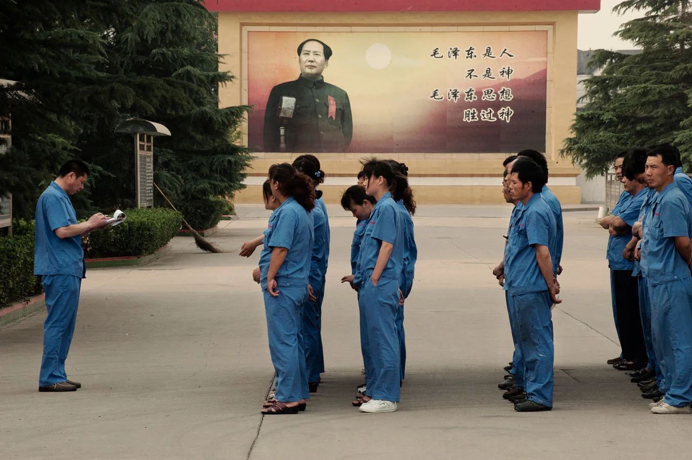 Workers standing up for the call before taking their shift at a factory in Nanjie. Behind them stands a large billboard with Chairman Mao and the slogan ""Mao Zedong is human being not a God, but Mao Zedong thought is greater than God".