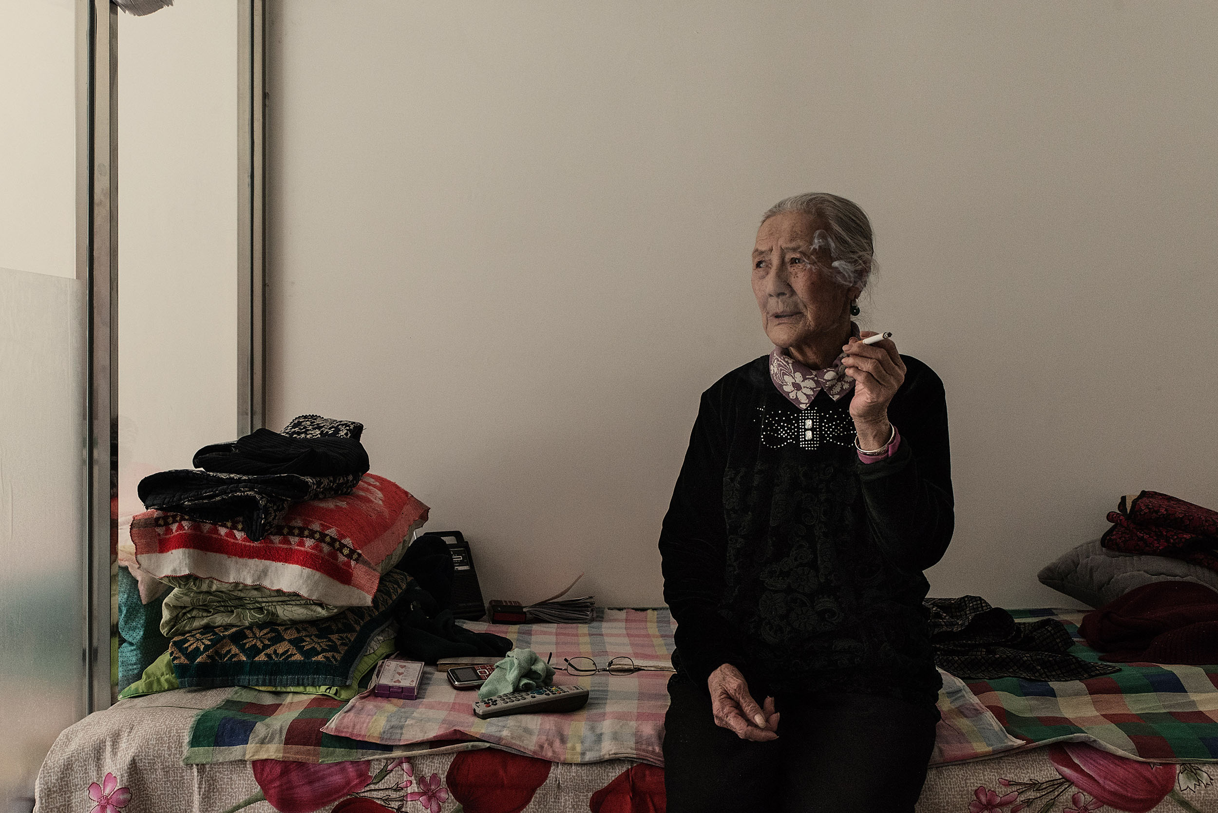 75 years old, Dushaocai sitting on her bed smoking, in Jarud Banner (Inner Mongolia). Dushaocai had to leave her grassland in Holingol due to pollution from a nearby aluminium mill. She was forced to resettle in an urban area, separated from her sons. Remembering her life on the grassland brings her to tears. Her best memory of, it is waking up before sunrise and cooking milk tea for her sons. Mongol nomads have been the first to be affected by forced resettlement, and wandering pastoralism a pillar of mongol culture and lifestyle, amost entirely disappeared in the 90s.