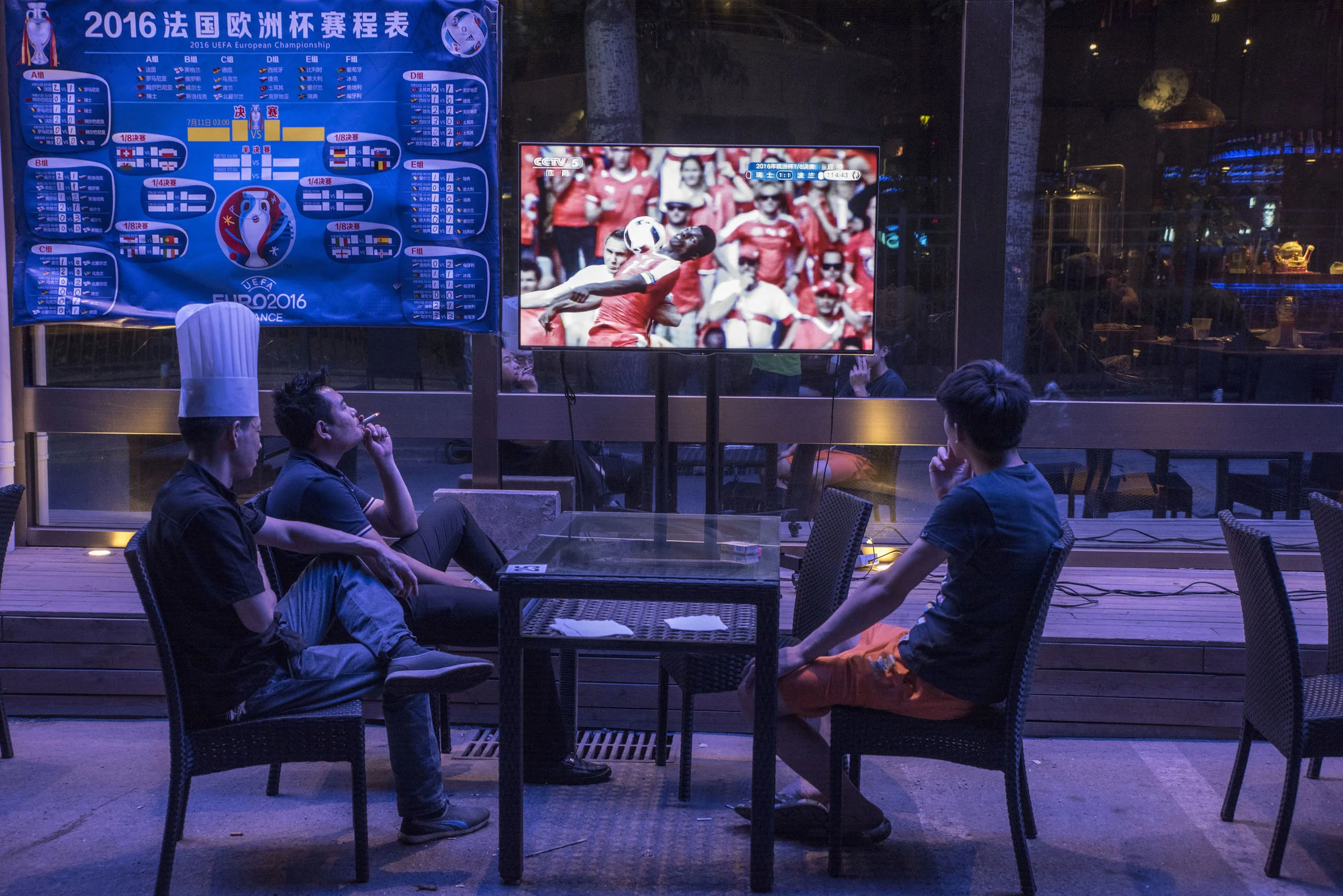 A cook and friends watching a game of the 2016 Eurocup, late at night on a street of Beijing. International football tournaments draw huge audiences in China, in spite of the absence of the National team which poor performances don't allow a presence in such games. The national men’s team recently placed 83rd in FIFA rankings, just ahead of the Faroe Islands.