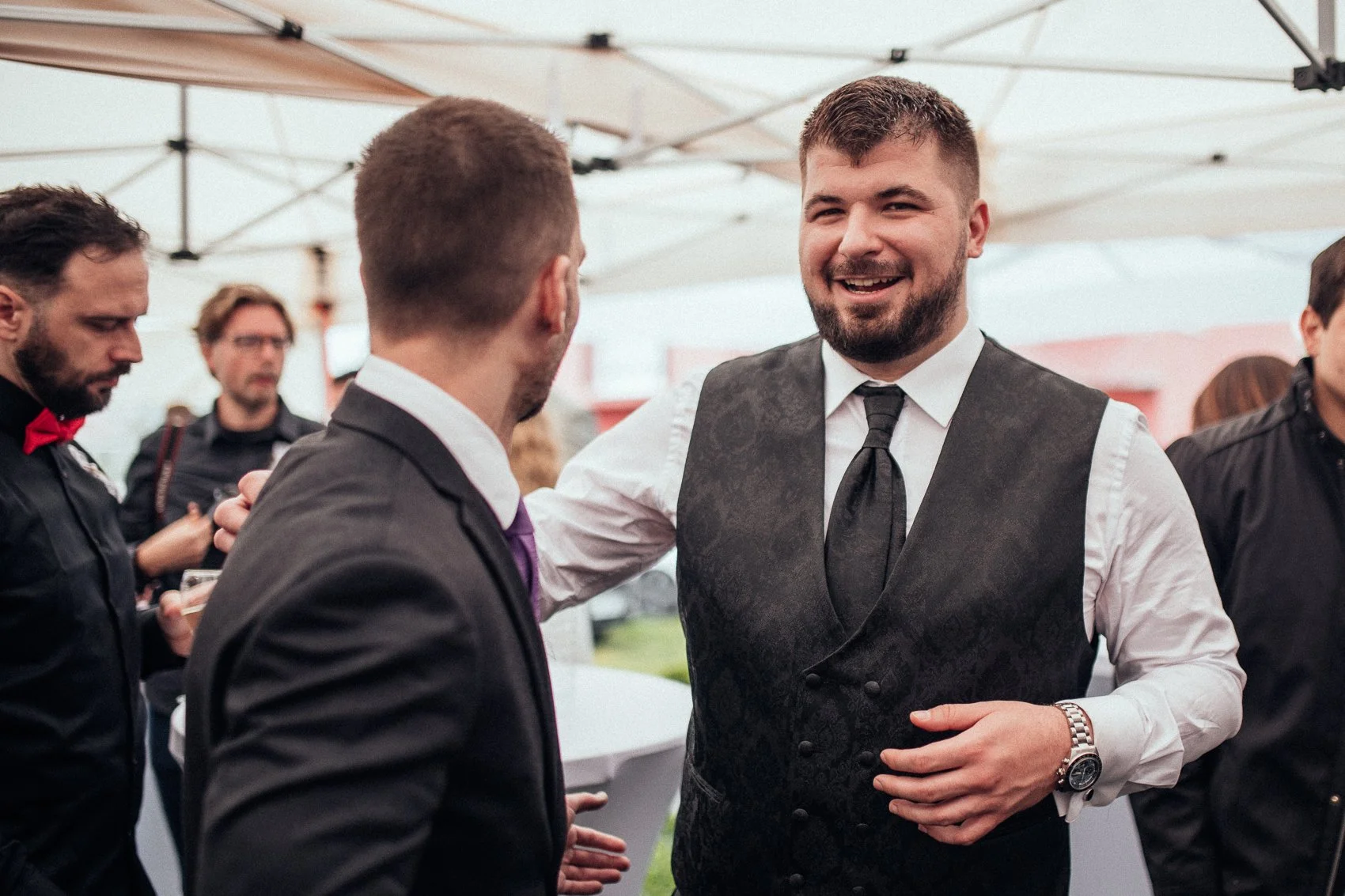 Group of men in formal attire, including vests, ties, and a red bow tie, socializing outdoors under a canopy.