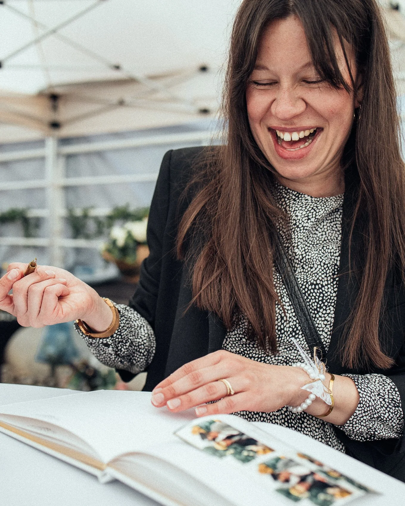A woman with long brown hair, smiling and laughing, sitting at a table with a photo album open in front of her. She is wearing a black blazer, a patterned black and white blouse, and has jewelry on her wrist and finger. The background shows a white o
