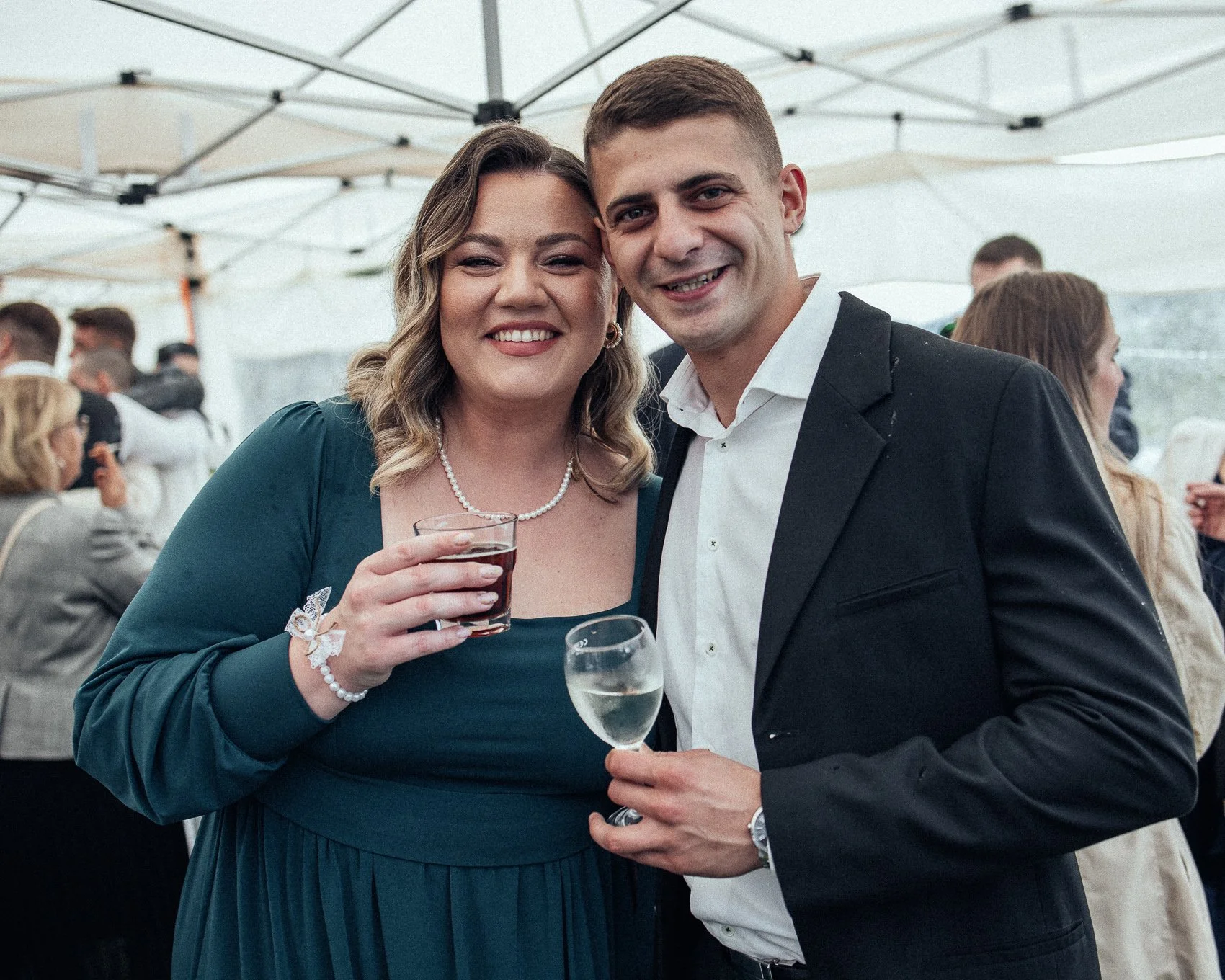 A smiling woman holding a glass of red drink and a smiling man holding a glass of white wine at an outdoor event under a canopy.