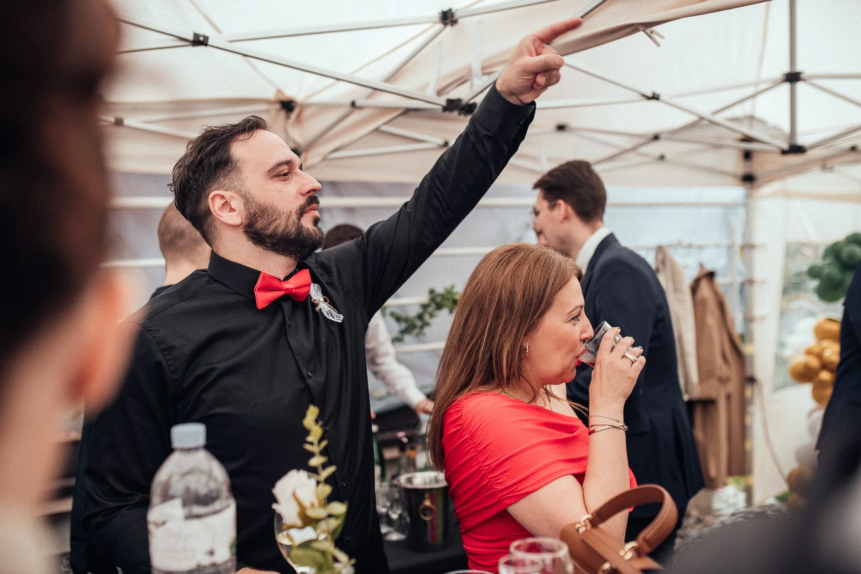 A man with a beard and black shirt with a red bow tie is pouring a drink at a social event. A woman with long red hair in a red dress is drinking from a glass nearby. The setting is under a canopy with other people in formal attire in the background.