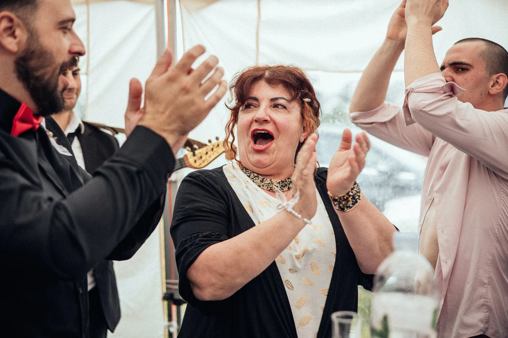 People celebrating at a party, clapping and smiling, with a woman in the center passionately engaging with others.
