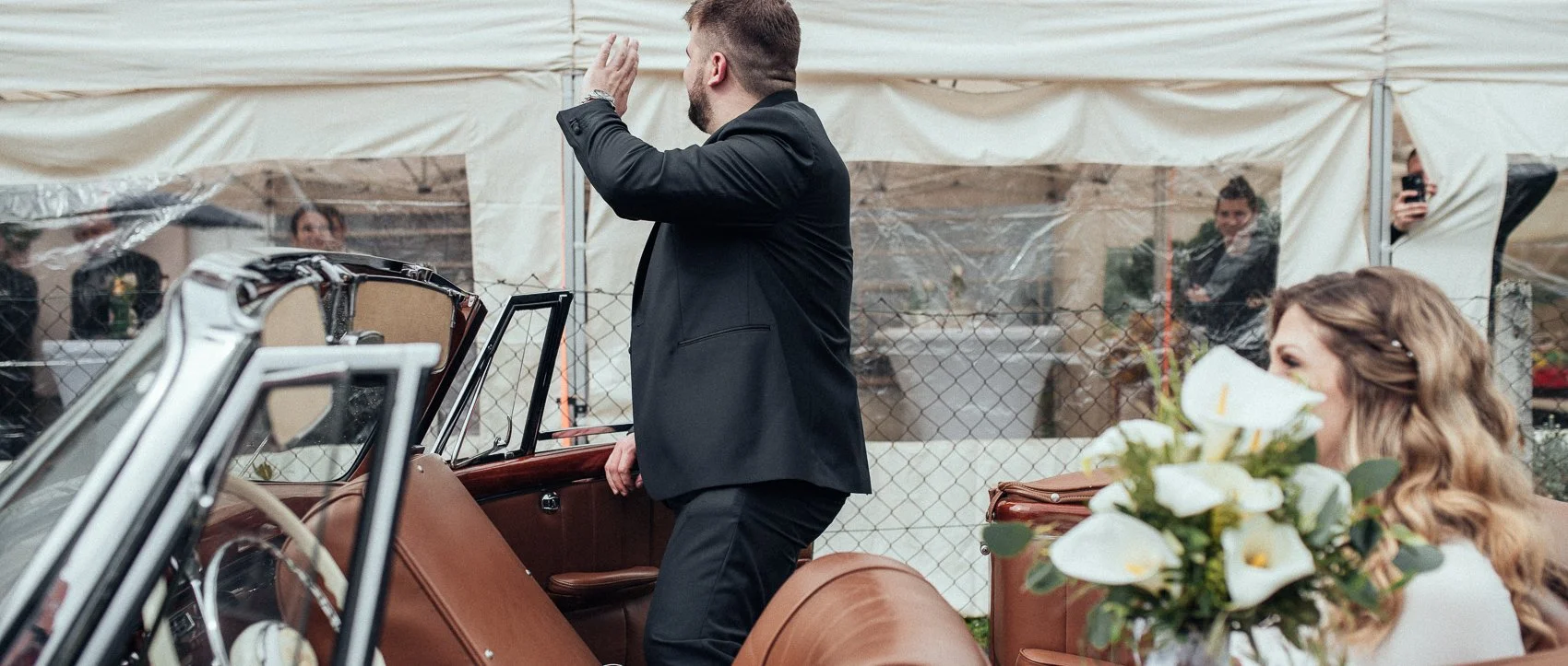 A man in a black suit giving a toast while standing beside a vintage convertible car, with a woman holding a bouquet of white flowers sitting nearby, in an outdoor setting with a tent and a chain-link fence.