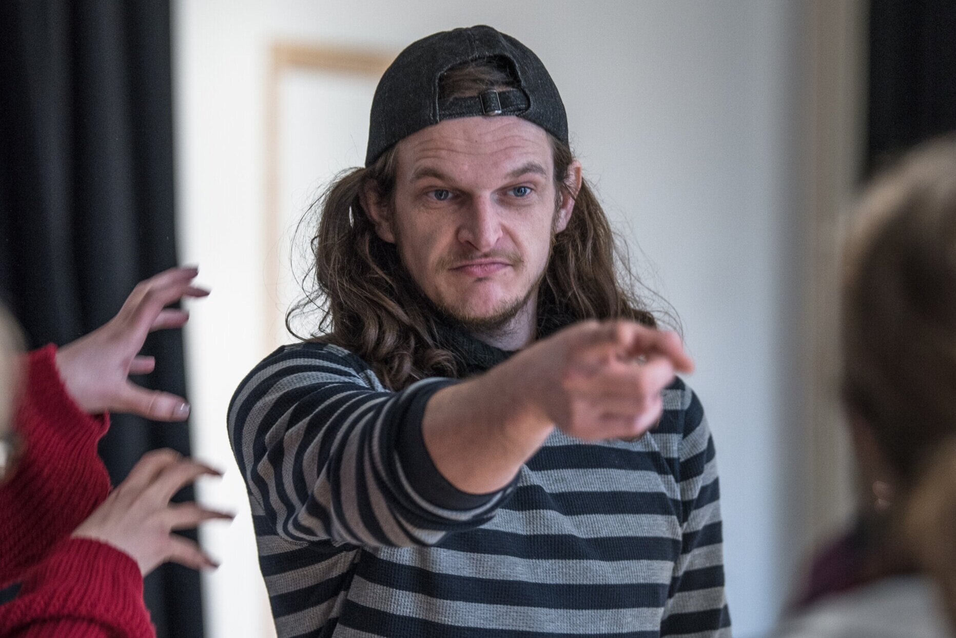  A white man with long hair wearing a baseball cap backwards and a stripey black and grey top points towards the person opposite him 