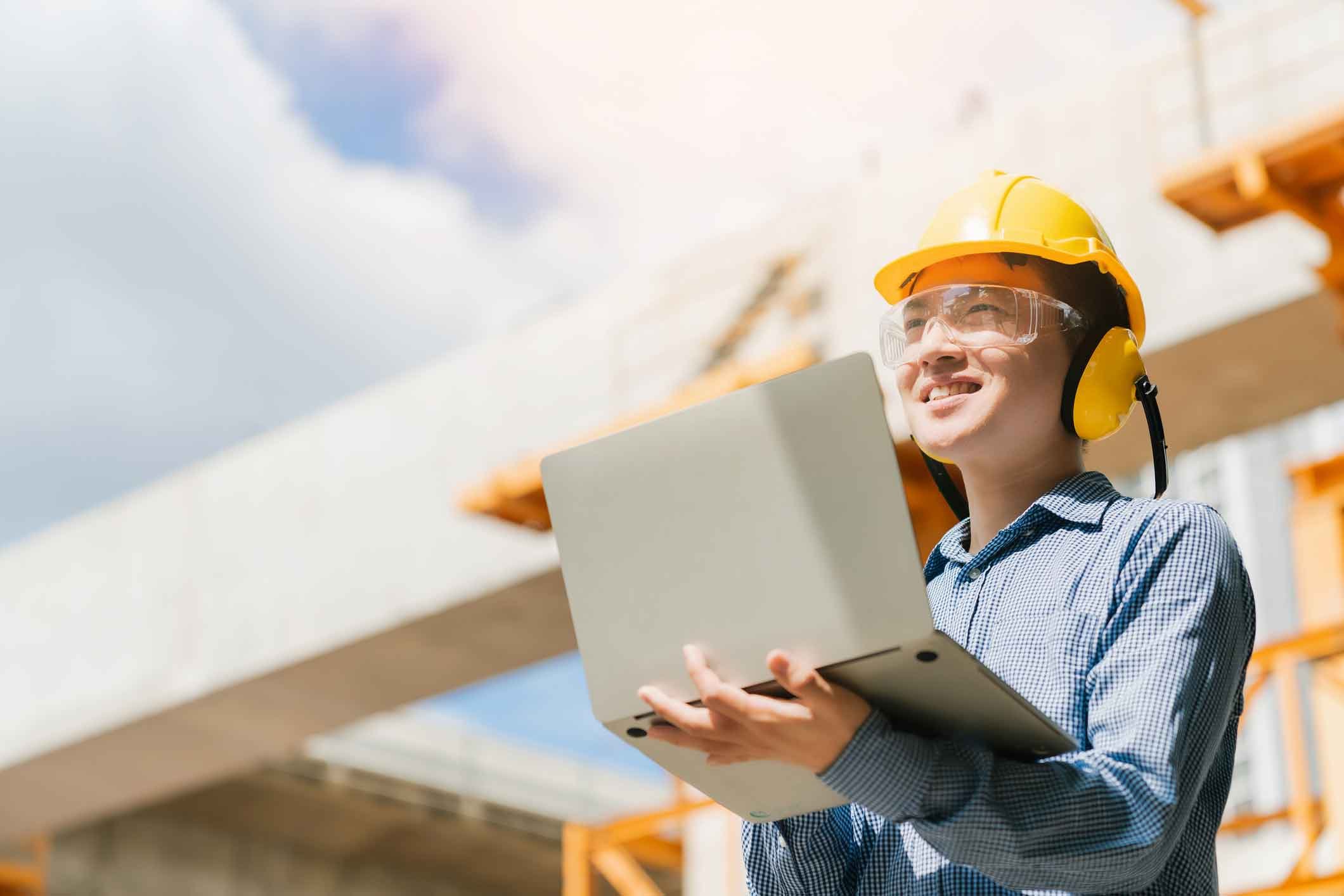 Employee wearing hard hat holding a laptop at a job site.
