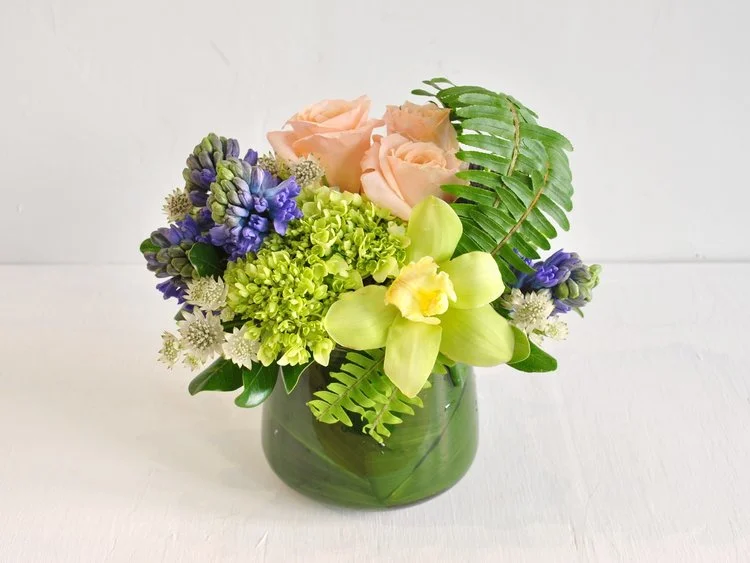 Soft playful tones of blue, peach and green bring about thoughts of the springtime thaw and picnics in a meadow. Fragrant hyacinth, shimmer roses, cream astrantia, chartreuse cymbium. hydrangea and sword ferns are collected in a rounded glass vase.