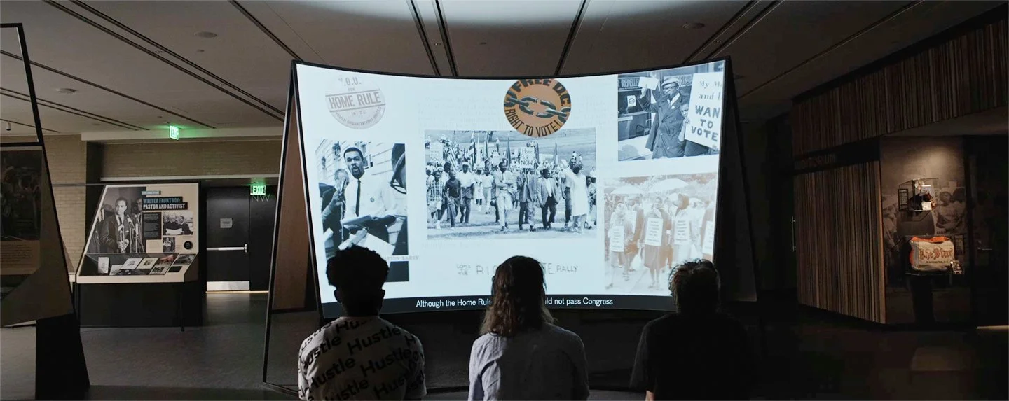 Three students, seated, watching a curved screen displaying a collage of ephemera from DC's Home Rule movement