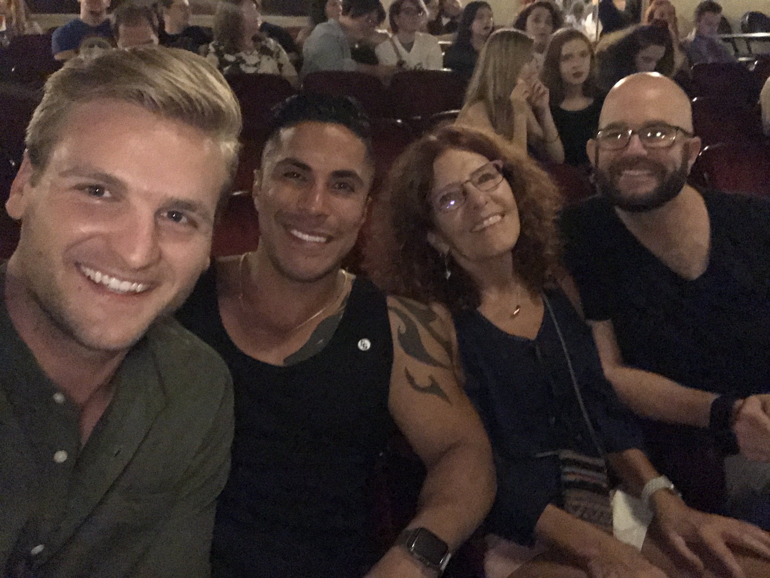 Un grupo de cuatro personas sonriendo en un lugar con asientos, posiblemente teatro o cine. Hay más personas en el fondo.