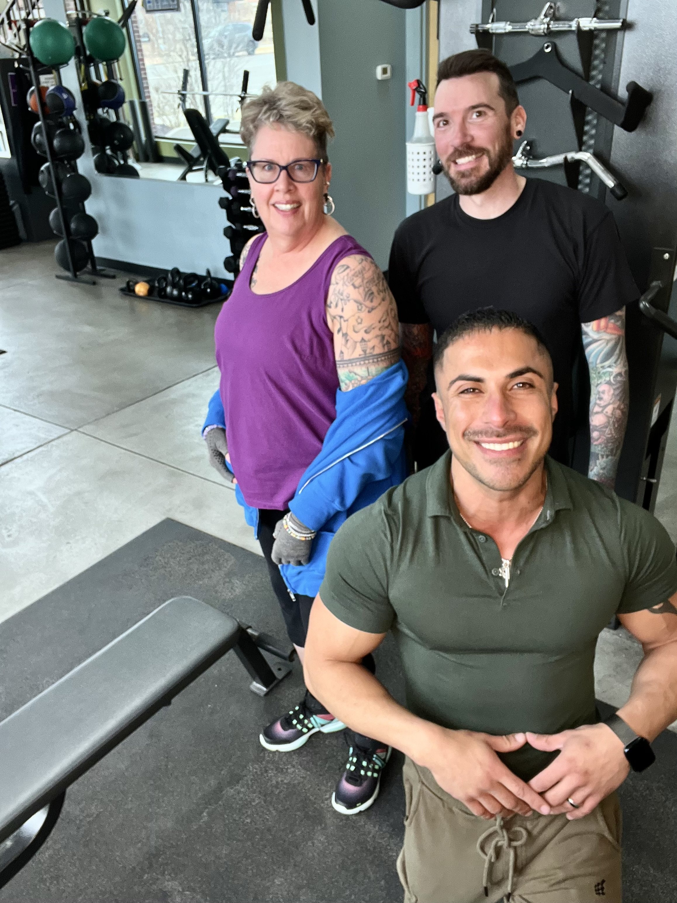 Tres personas sonriendo en un gimnasio, con equipo de entrenamiento en el fondo y una banca de ejercicios en primer plano.