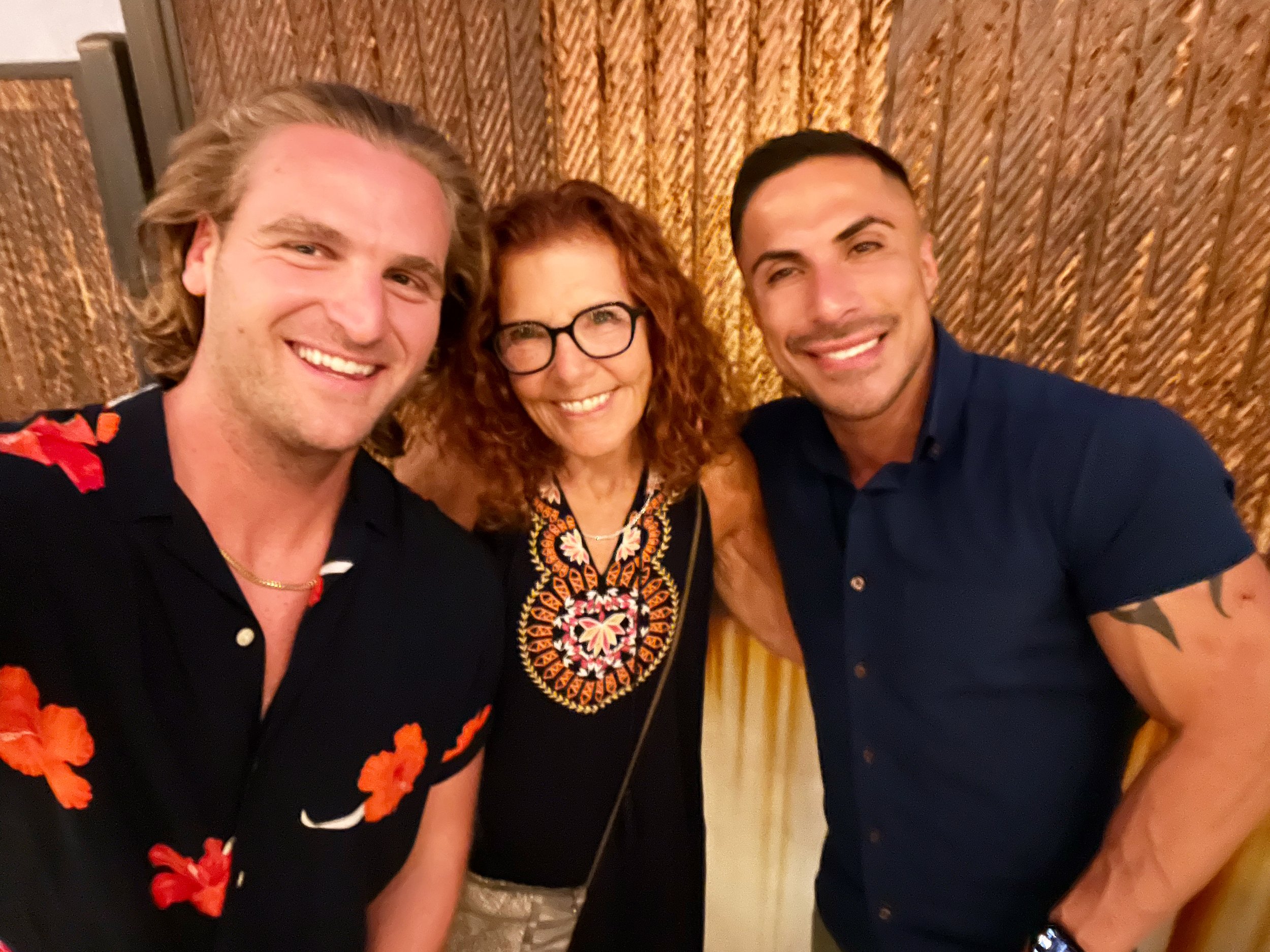 Tres personas sonriendo frente a una pared decorativa. Una mujer en el centro con gafas y vestido estampado, flanqueada por dos hombres que visten camisas informales. Ambos hombres tienen sonrisas amplias y distintas características físicas.