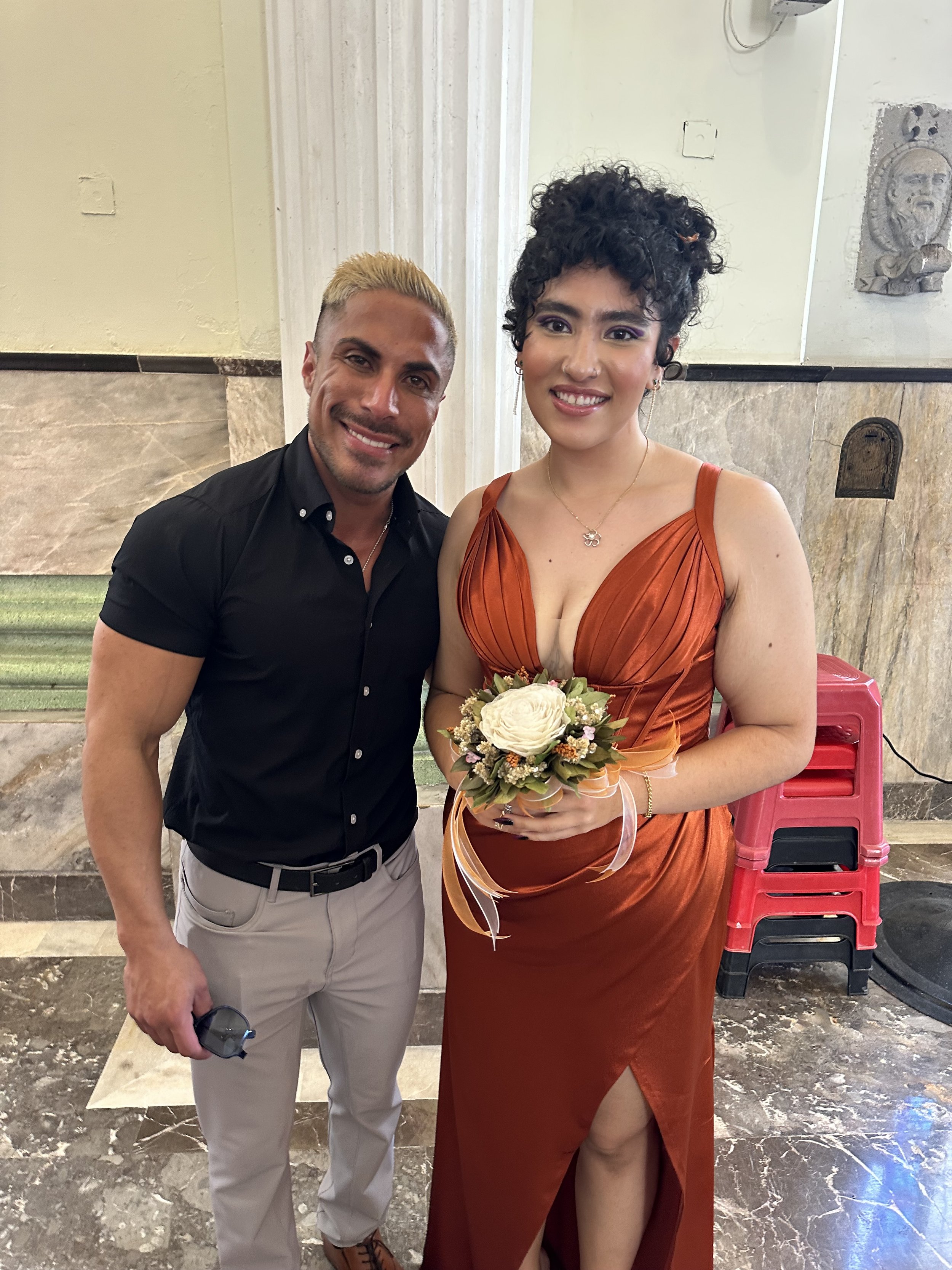 Dos personas, una mujer con un vestido naranja sosteniendo un ramo de flores, y un hombre con camisa negra y pantalón claro, posando juntos en un interior con columnas y decoración clásica.