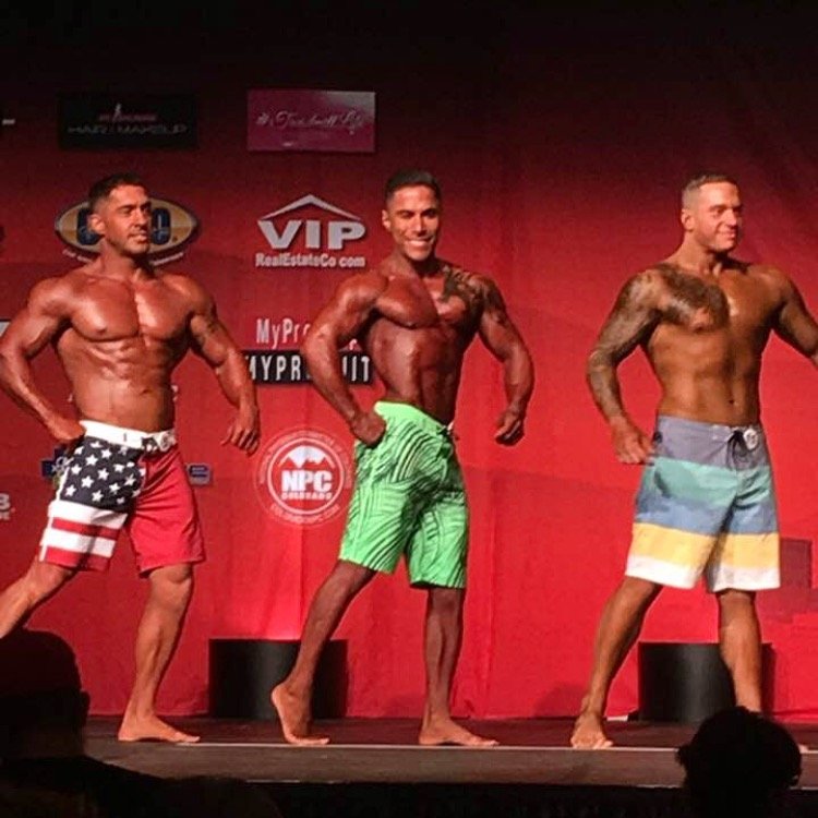 Tres culturistas posan en el escenario de una competición, con pantalones cortos de colores y un fondo rojo con logotipos de patrocinadores.