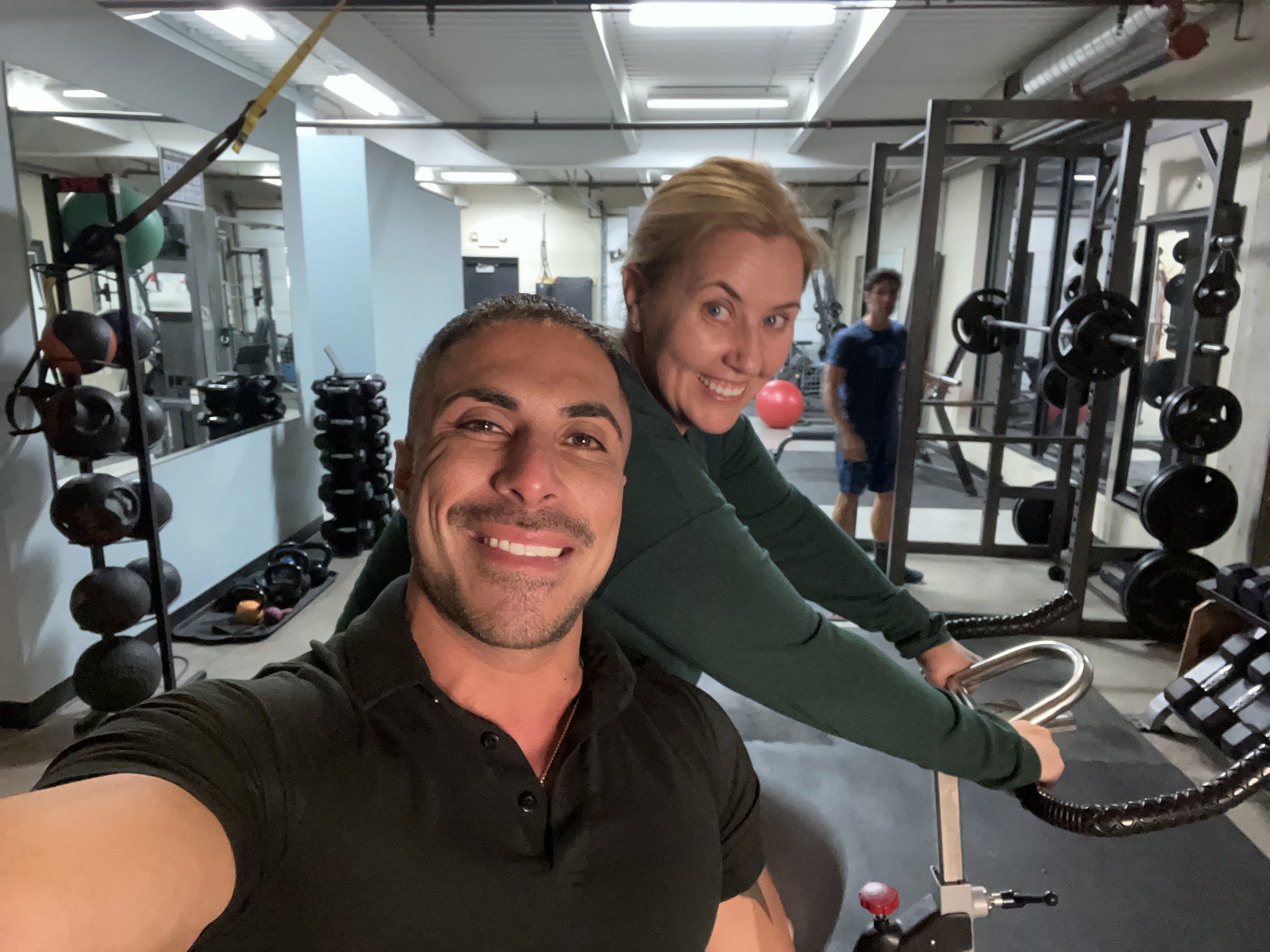 Dos personas sonrientes en un gimnasio con equipo de ejercicio y pesas en el fondo.