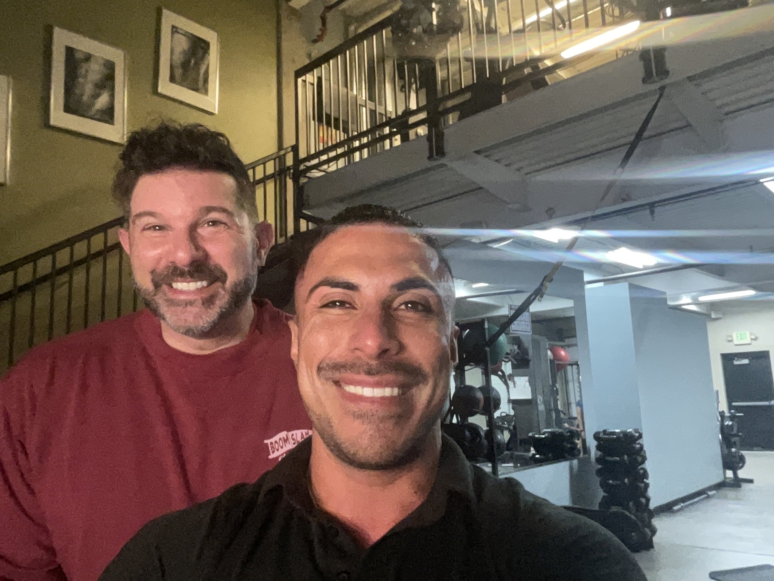 Dos hombres sonrientes en un gimnasio con equipos de fitness al fondo.