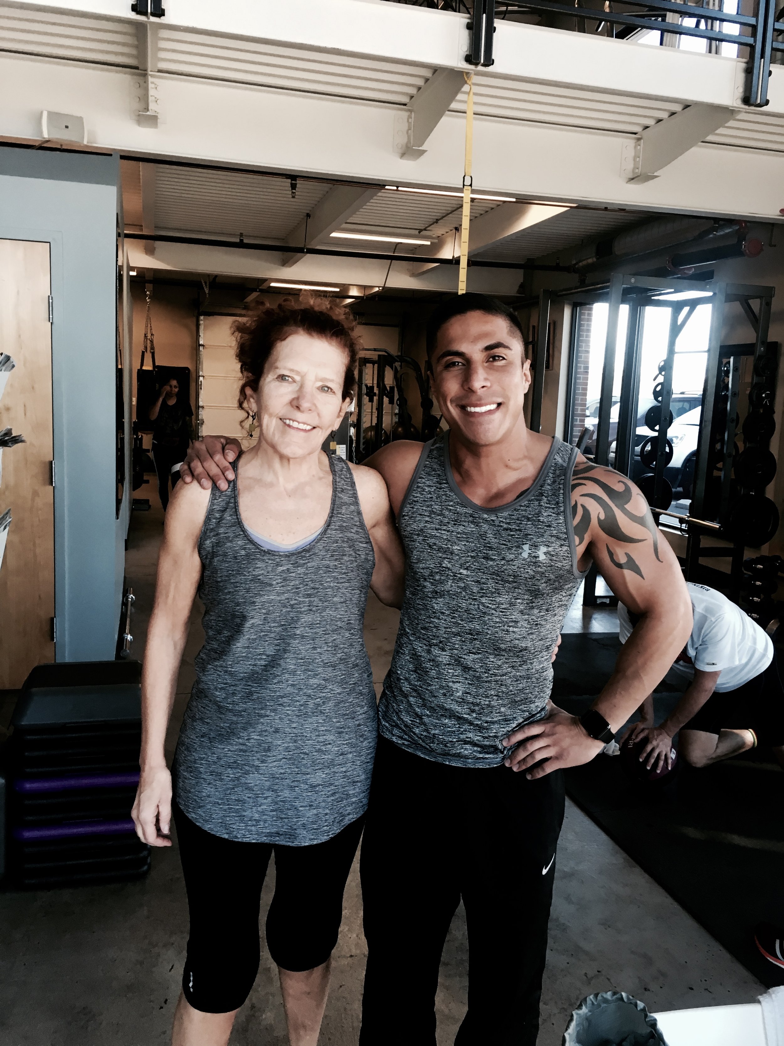 Dos personas en ropa deportiva posando en un gimnasio con equipo de entrenamiento en el fondo.