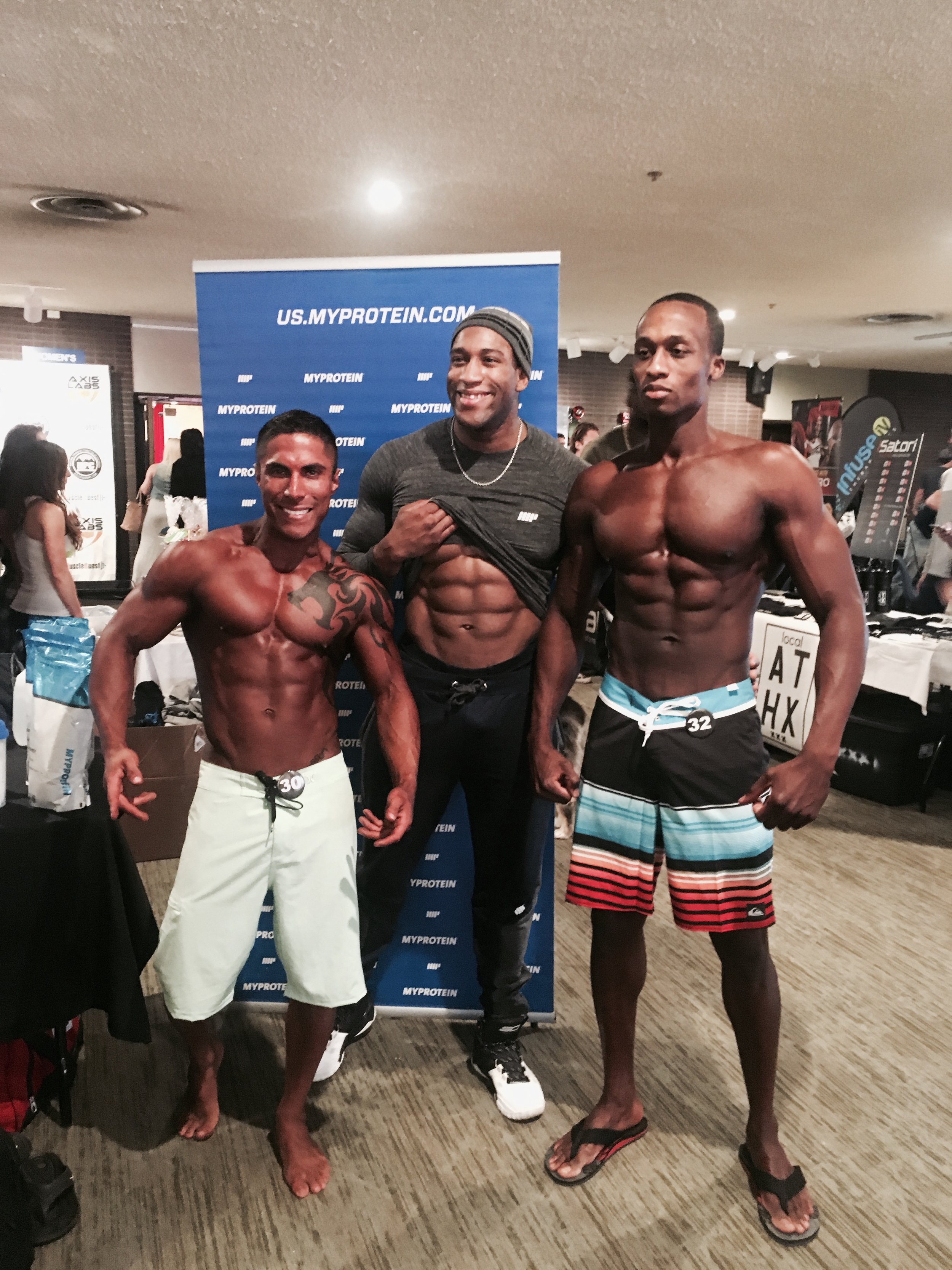 Tres hombres musculosos posan frente a una pancarta de MyProtein. Están en un evento relacionado con el fitness. Dos de ellos visten pantalones cortos y el otro un pantalón deportivo.