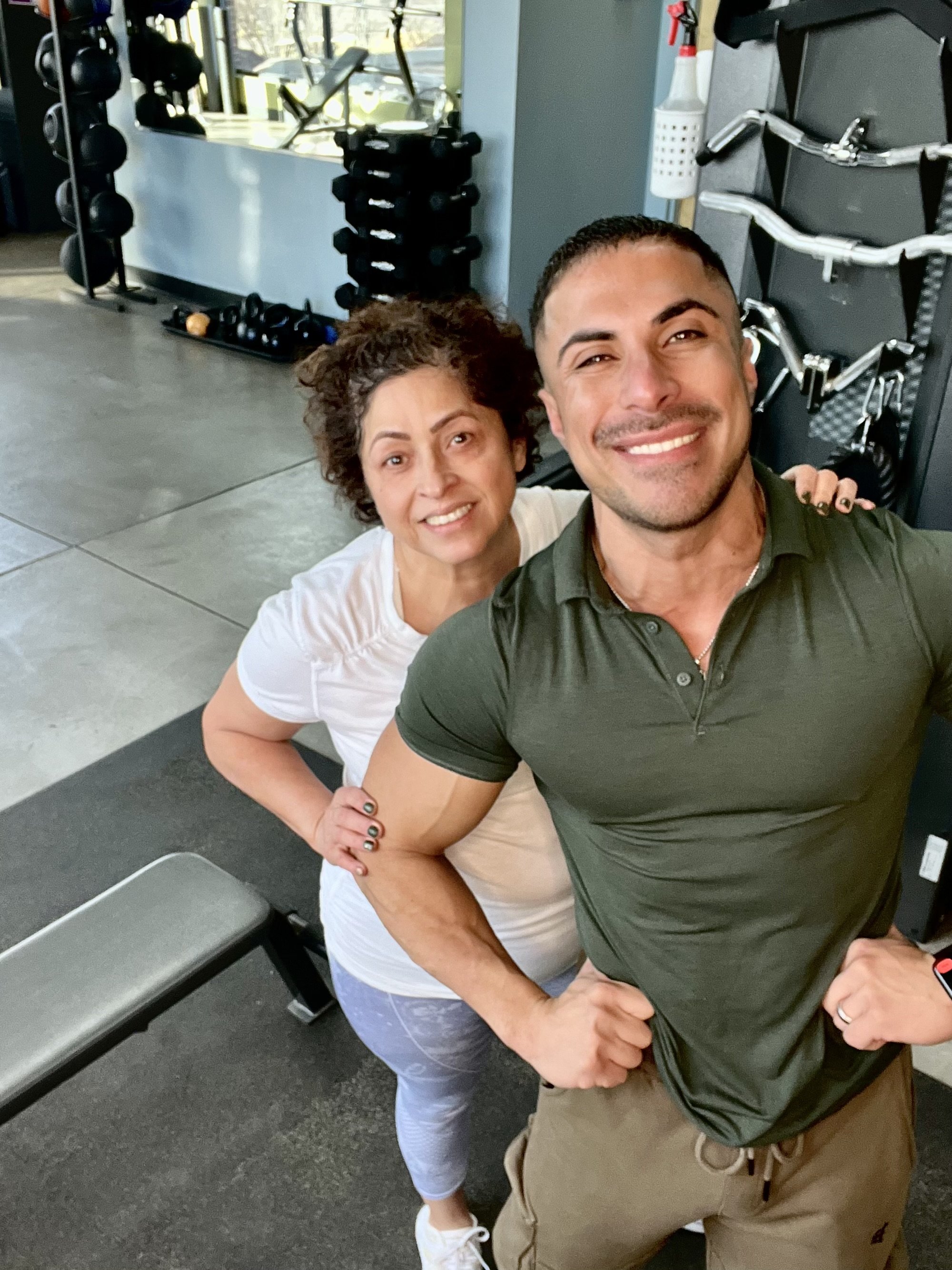 Dos personas sonriendo en un gimnasio, con equipo de ejercicio en el fondo.