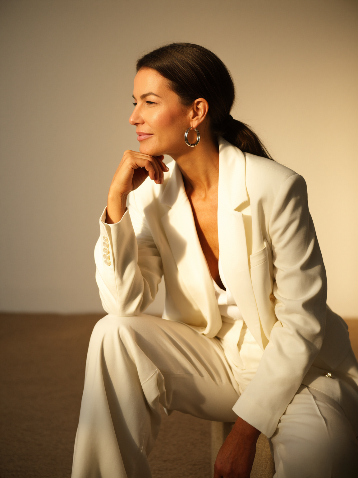 A woman in a white suit sitting with her hand on her chin, looking to the side, in a warmly lit setting.