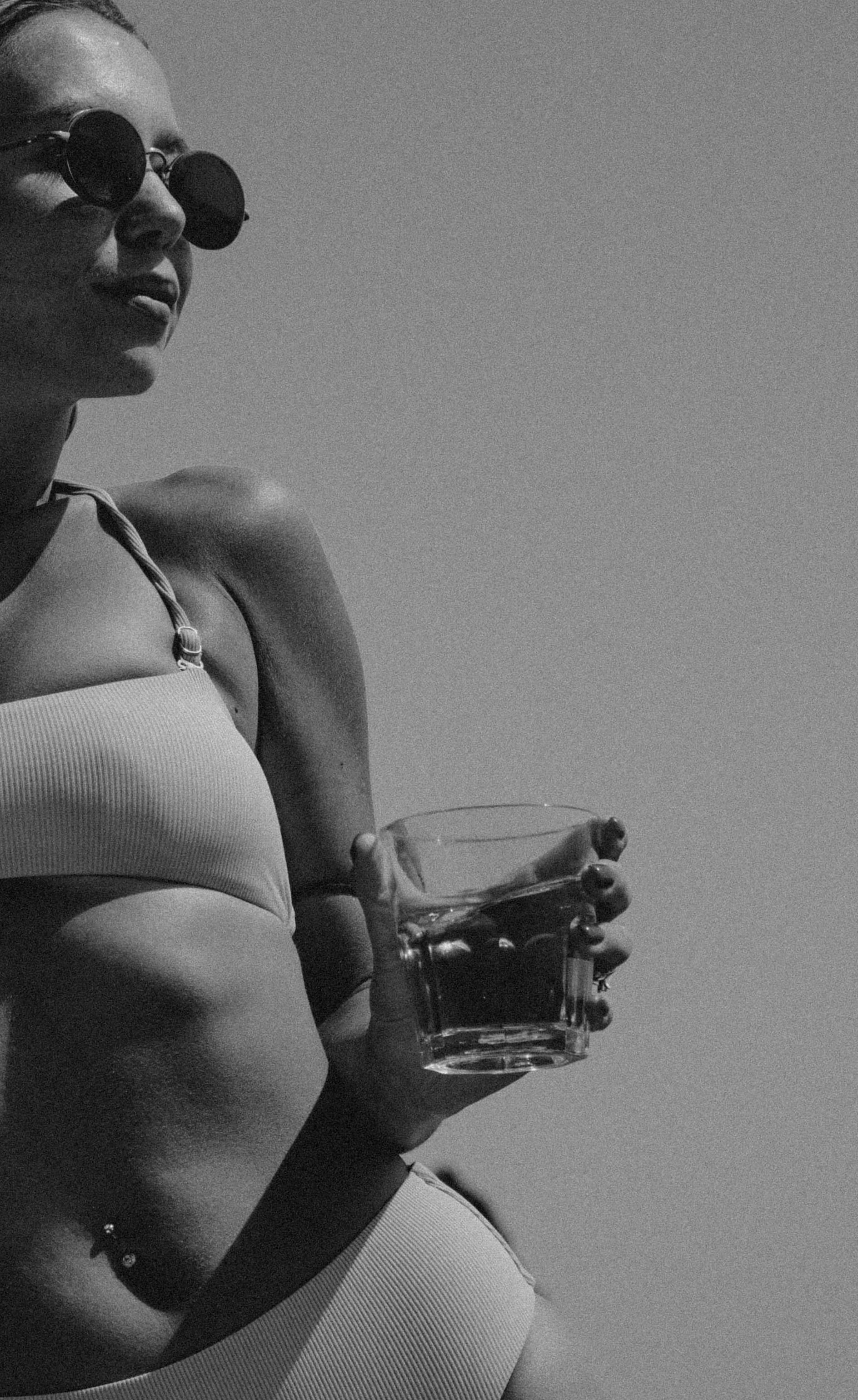A woman wearing sunglasses and a bikini holding a glass of water, relaxing outdoors.